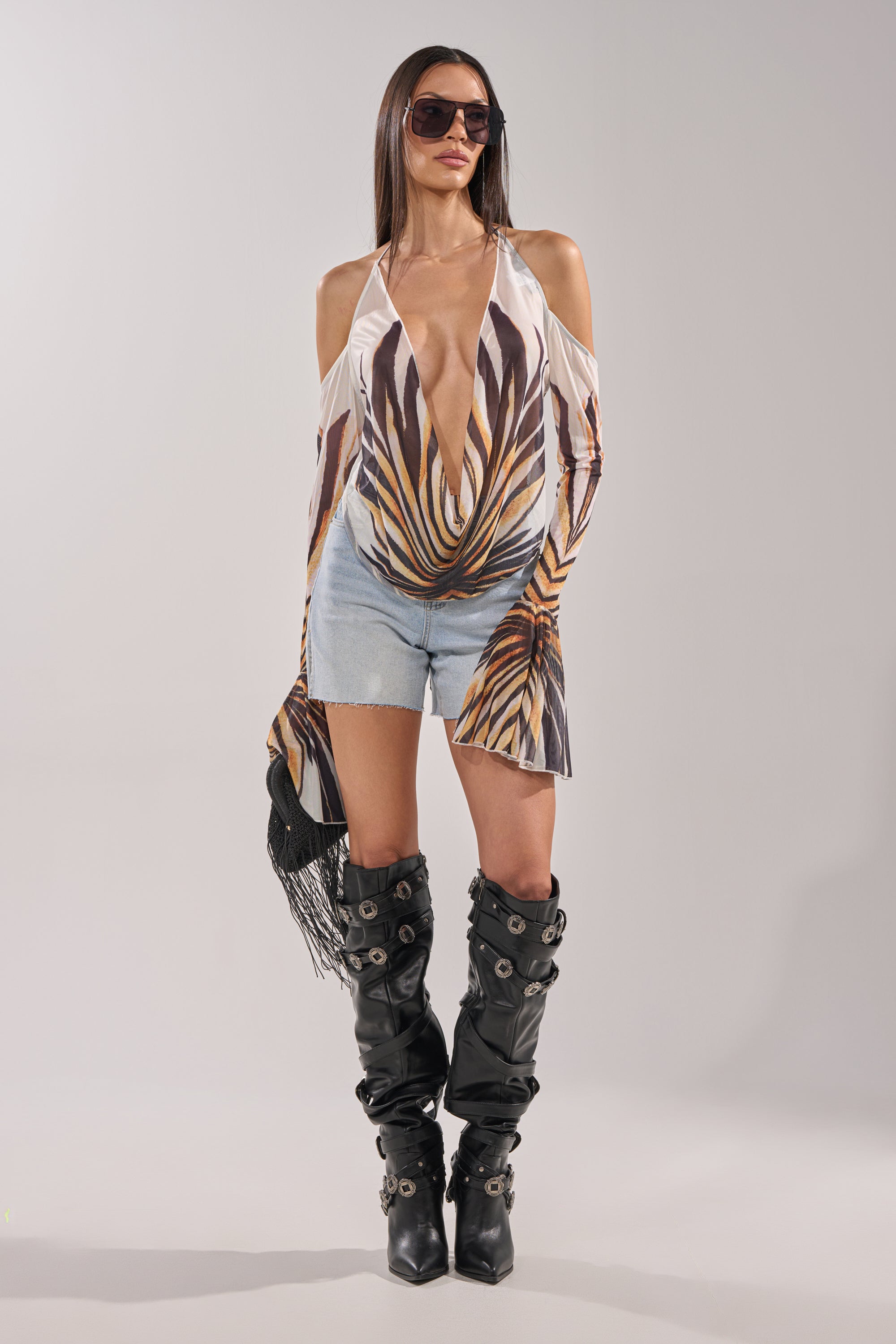 A woman stands confidently against a plain background wearing sunglasses, the PRIMAL SPIRIT BLOUSE with a deep V-neck and leaf pattern, light blue denim shorts, and black knee-high boots with metallic details. 6