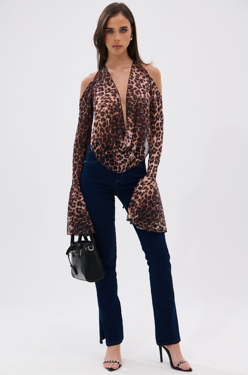 A woman stands against a white background wearing the CHEETAH WORLD BLOUSE with leopard print, off-the-shoulder and open back design, flared cuffs, paired with dark blue jeans, black high-heeled sandals, and a small black handbag.