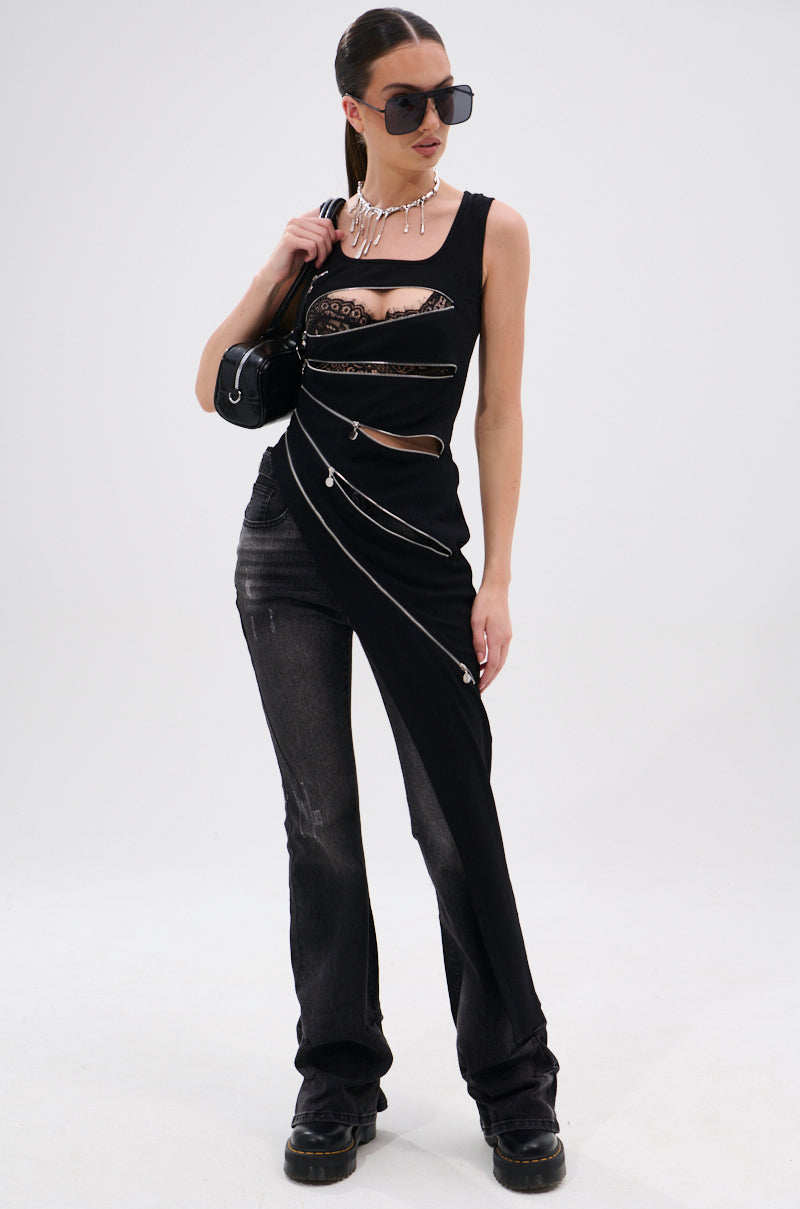 A woman in black sunglasses, a statement necklace, and the AKIRA Label TOO GOOD FOR YOU BLOUSE WITH ZIPPERS with lace details and edgy zippers. Her asymmetrical tank features one pant leg, and she carries a black handbag on a white background.