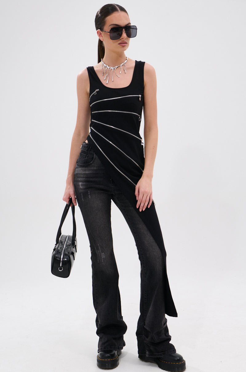 A person wearing oversized sunglasses, a necklace, the TOO GOOD FOR YOU BLOUSE WITH ZIPPERS with diagonal white lines, black distressed jeans with metallic zippers, black boots, and holding a black handbag stands against a white background.
