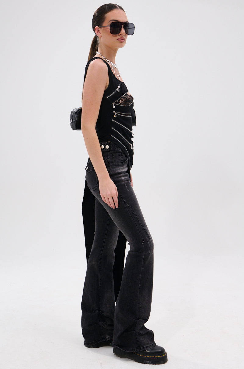 A woman in sunglasses, the TOO GOOD FOR YOU BLOUSE WITH ZIPPERS—a black asymmetrical tank with metallic details—and dark flared jeans and black boots stands against a white background. Her hair is styled in a sleek ponytail.