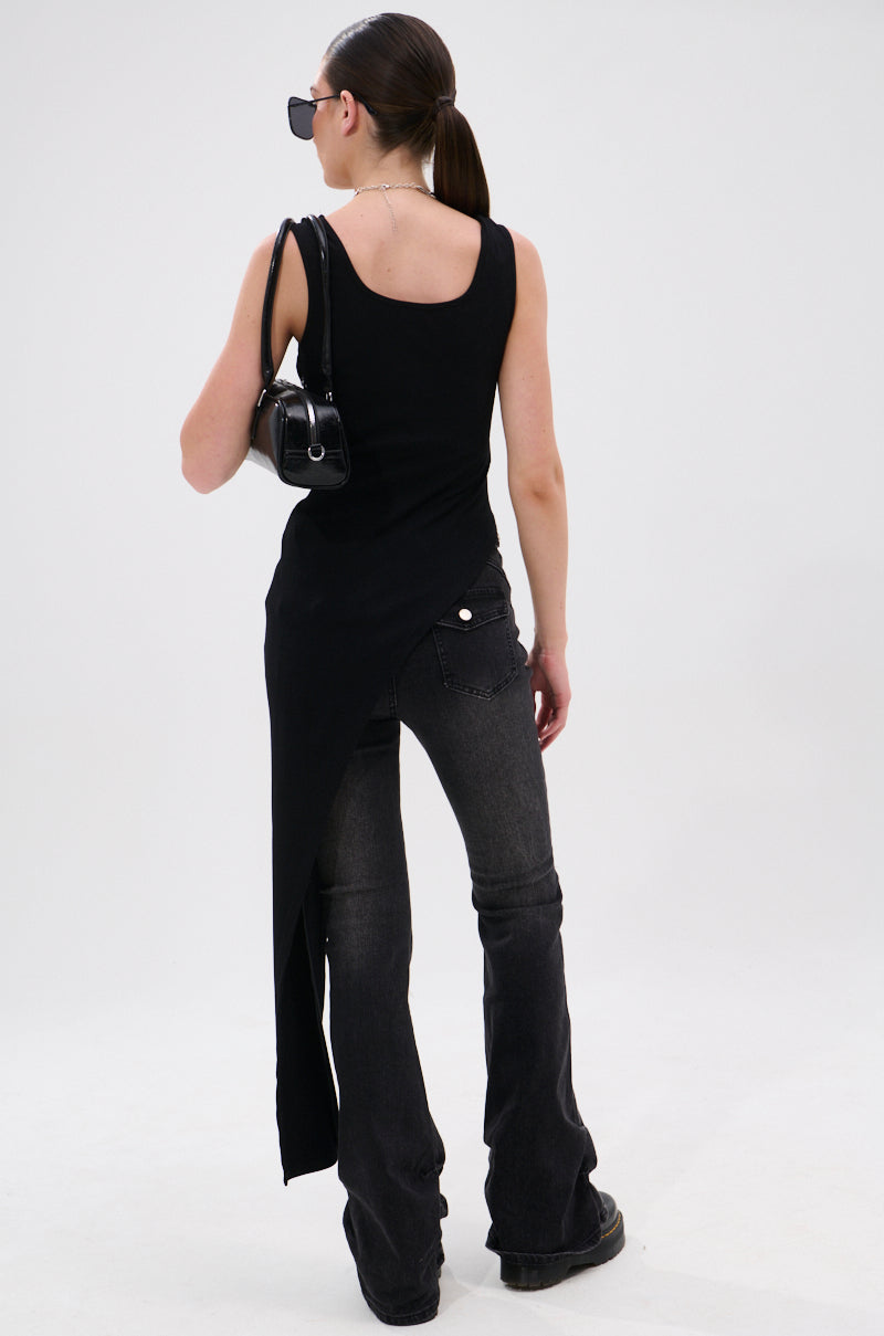 A woman stands facing away, wearing the TOO GOOD FOR YOU BLOUSE WITH ZIPPERS, dark flared jeans with metallic zippers, black shoes, sunglasses, and a black shoulder bag. Her hair is in a low ponytail against a plain white background.