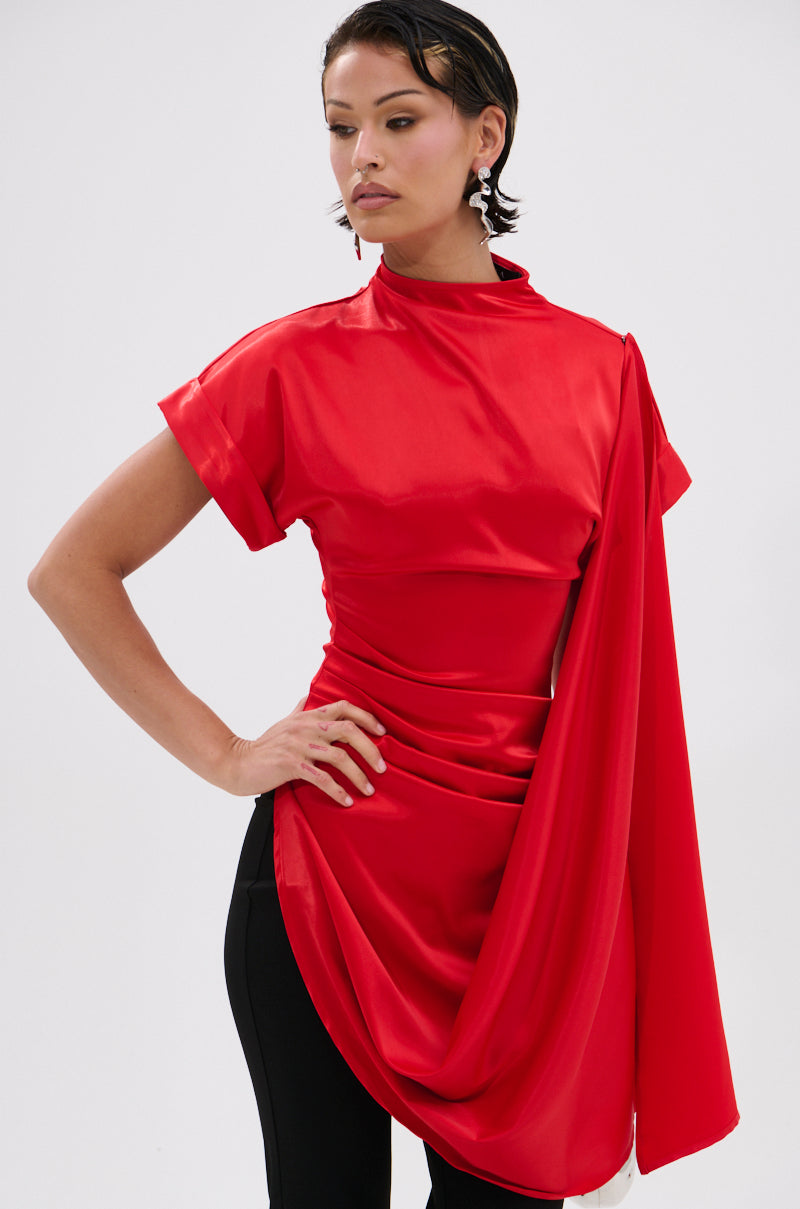 A person poses confidently in the bright red HILLARY SATIN BLOUSE, featuring an asymmetrical cut, high neckline, and short sleeves, paired with black pants. They accessorize with statement earrings and have short slicked-back hair against a plain white background.