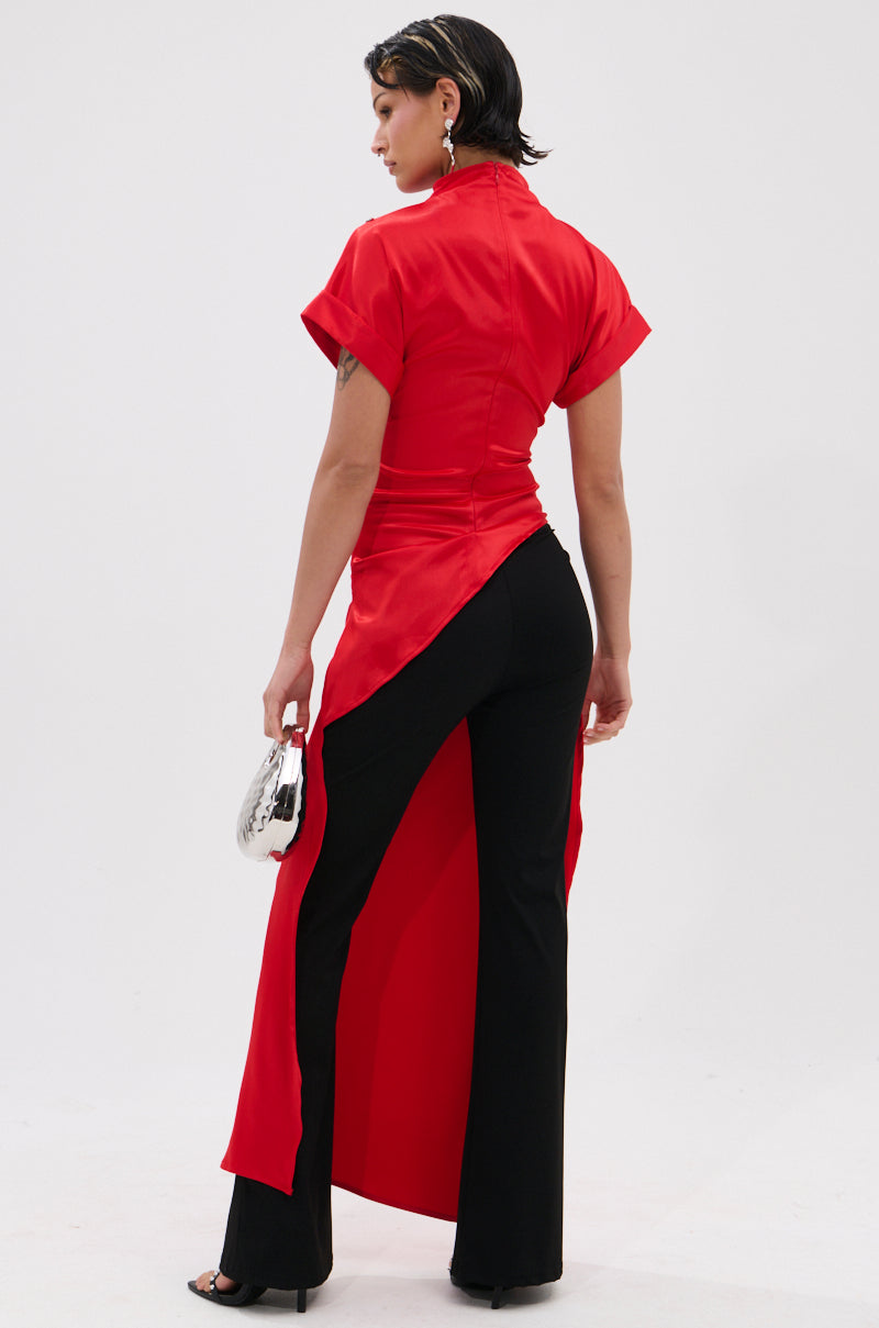 A person stands facing away, wearing the HILLARY SATIN BLOUSE in red with short sleeves and an asymmetrical high-slit hem, paired with black pants, heels, and a silver handbag—an elegant evening look against a white background.