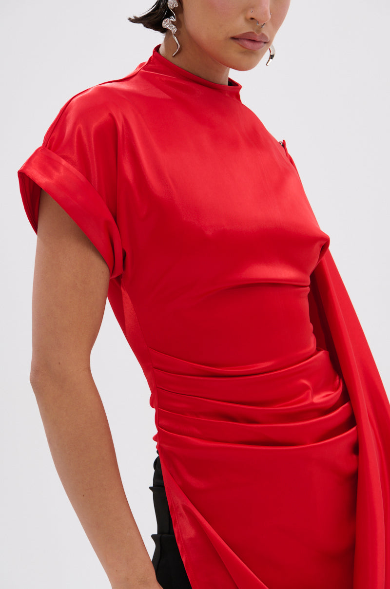 A person with short hair and geometric silver earrings wears the HILLARY SATIN BLOUSE—a shiny, bright red satin top with short sleeves, draped fabric, and rolled cuffs. The background is plain white.
