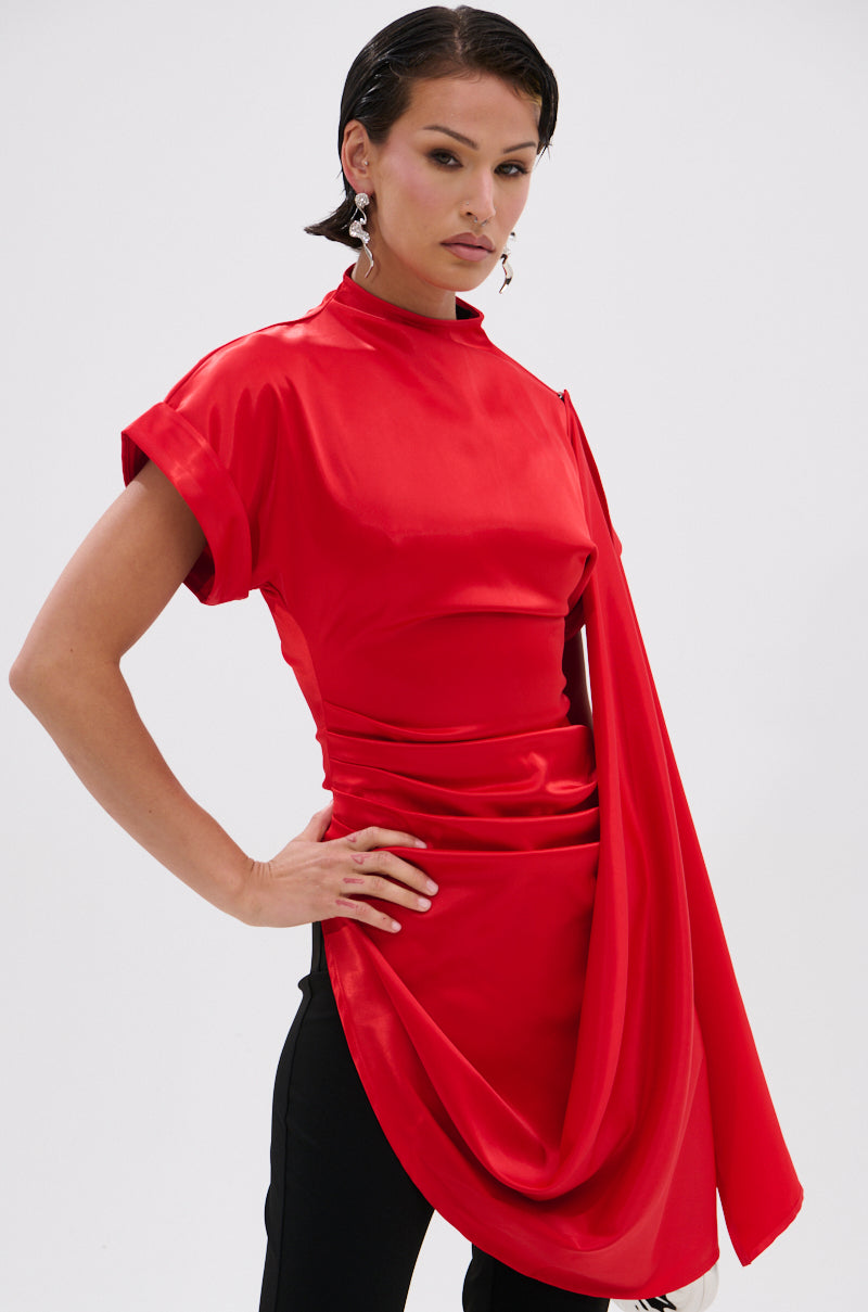 A woman with short dark hair wears the HILLARY SATIN BLOUSE in vibrant red, featuring draped detailing and short sleeves, paired with black pants. She stands confidently against a plain white background.