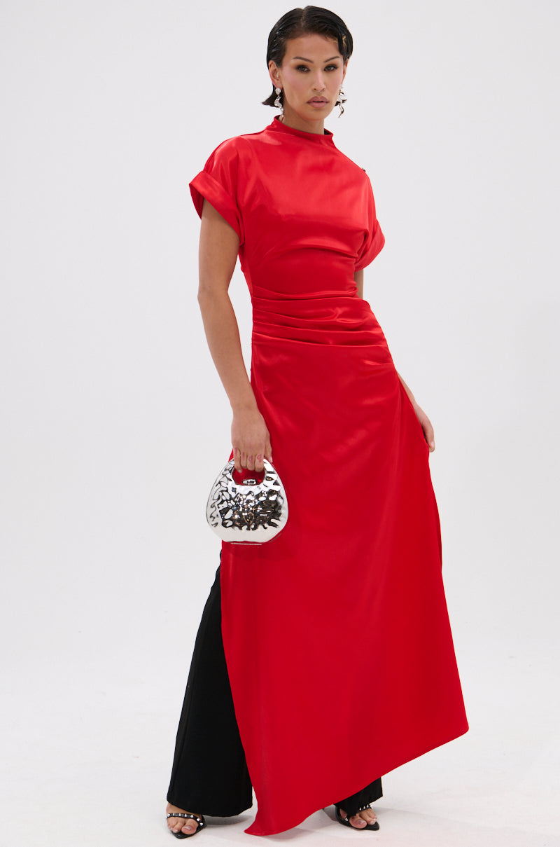 A person stands confidently in the HILLARY SATIN BLOUSE, a bright red asymmetrical top styled with black pants, black heeled sandals, and a shiny silver handbag against a plain white background for an elevated eveningwear look.