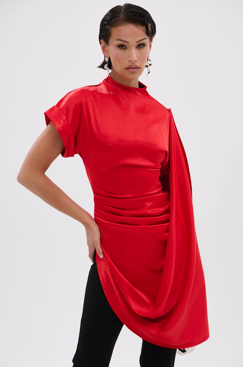 A person models the HILLARY SATIN BLOUSE—a bright red, asymmetrical satin top with draped details and short sleeves—paired with black pants against a plain white background, creating a chic contemporary evening look.