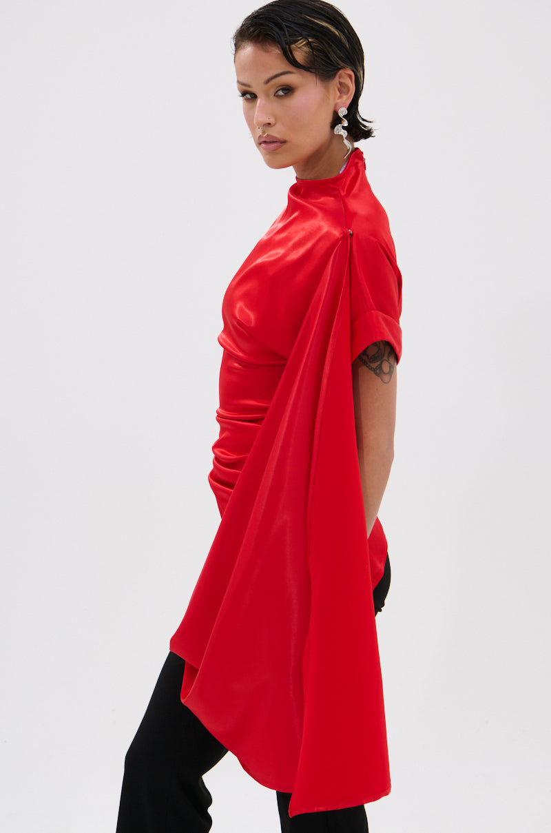 A person with short dark hair wears the HILLARY SATIN BLOUSE, a shiny red asymmetrical satin top with a dramatic draped sleeve, paired with black pants and posed against a plain white background.