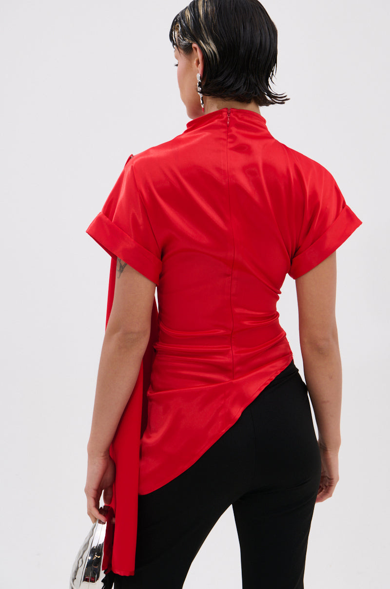 A person with short dark hair, seen from behind, wears the HILLARY SATIN BLOUSE in shiny red satin with an asymmetrical hem and black pants, holding a small clear handbag against a plain light background.
