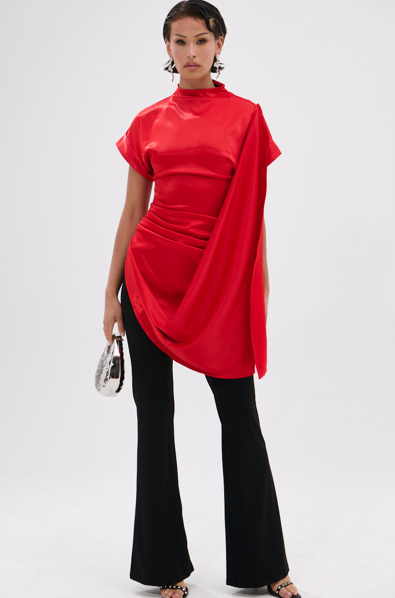 A person models the HILLARY SATIN BLOUSE—an asymmetrical shiny red satin top with draped fabric—styled with black flared pants, strappy heels, and a small silver bag against a white background for chic eveningwear.