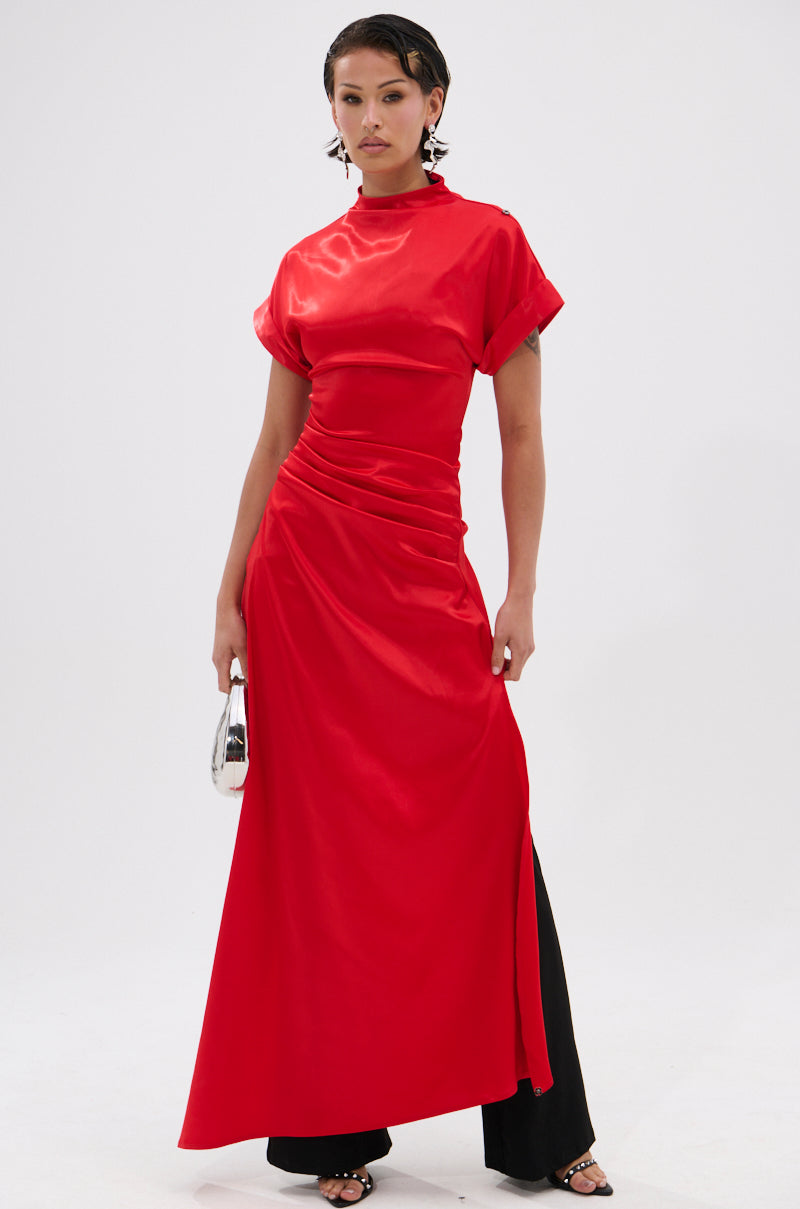 A woman models the HILLARY SATIN BLOUSE in shiny, bright red satin with short sleeves, a high neckline, and ruched details. She pairs it with black pants and a silver clutch against a white background—ideal for modern eveningwear.