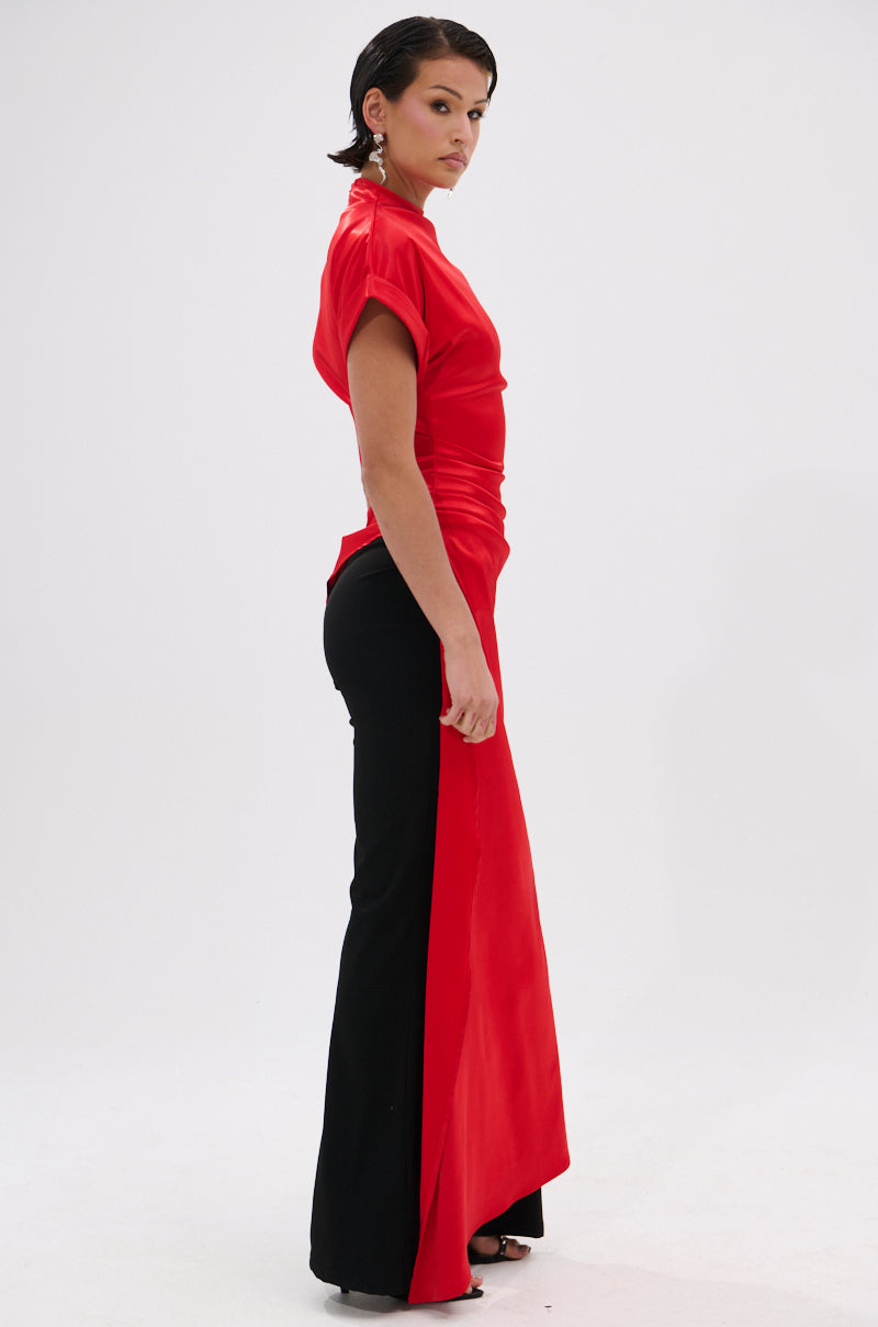 A woman poses sideways in the HILLARY SATIN BLOUSE, featuring an asymmetrical cut and long red sash, styled for eveningwear with black flared pants and heels, set against a plain white background.