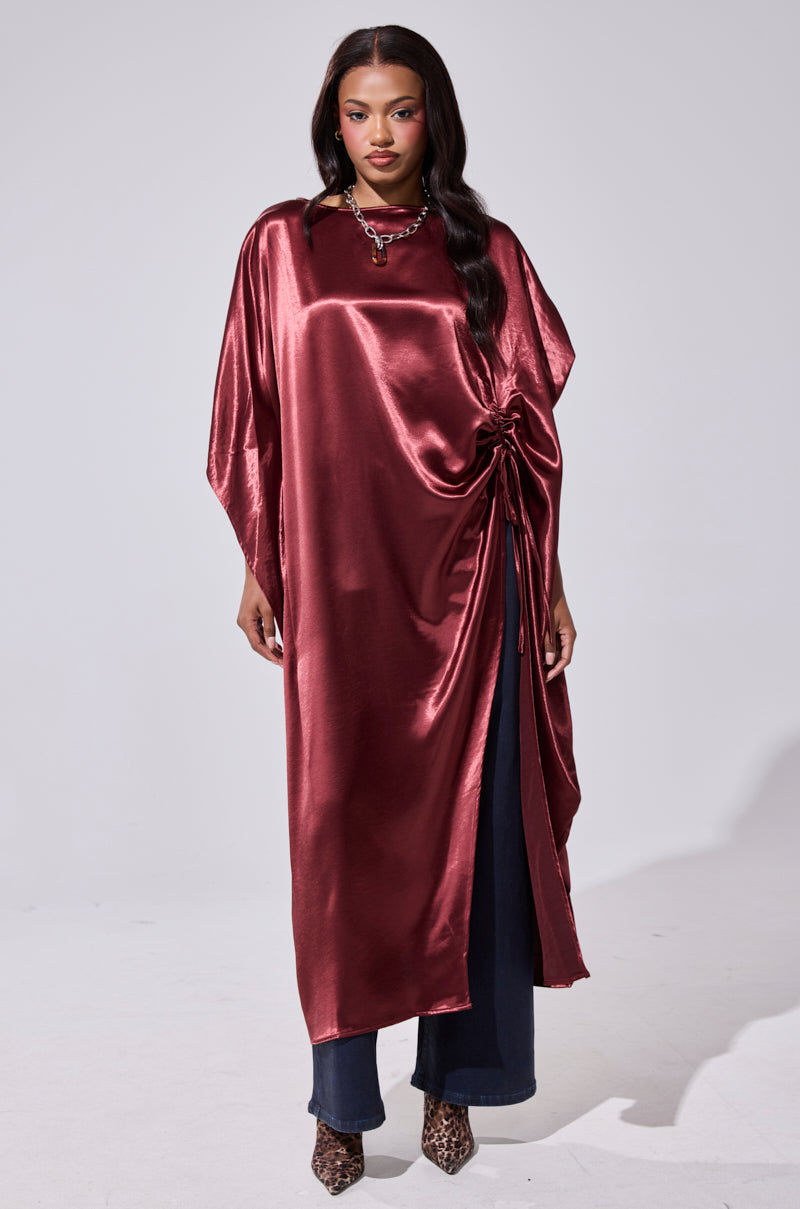 A woman stands facing forward, wearing the CLARA SIDE RUCHED BLOUSE in shiny burgundy satin with an asymmetrical hem and side-gathered knot, paired with dark pants, a statement necklace, and leopard-print heels. 1