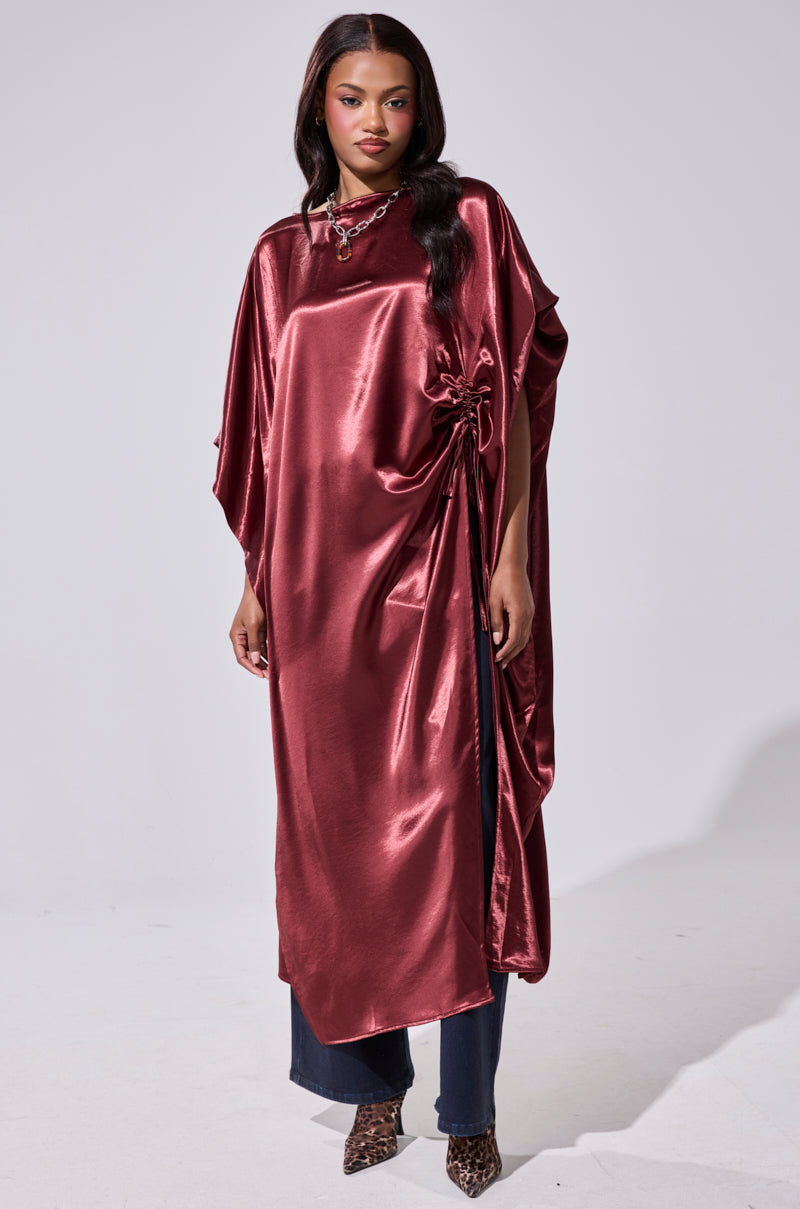 A woman models the CLARA SIDE RUCHED BLOUSE, a shiny maroon oversized satin top with side gathering. She pairs it with dark pants, a necklace, and animal-print shoes, and wears her long dark hair loose. 2