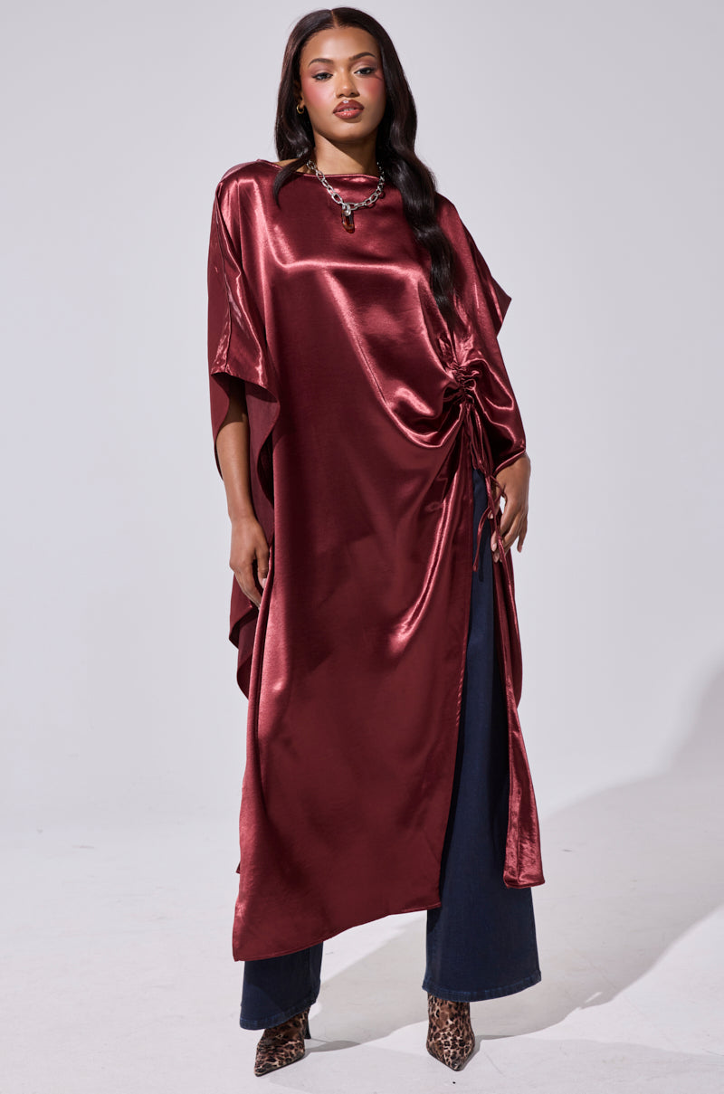 A woman poses confidently in the CLARA SIDE RUCHED BLOUSE, a shiny dark red satin top with side ruching and an asymmetrical hem, paired with flared dark blue pants, a necklace, and leopard print heels against a plain background. 0