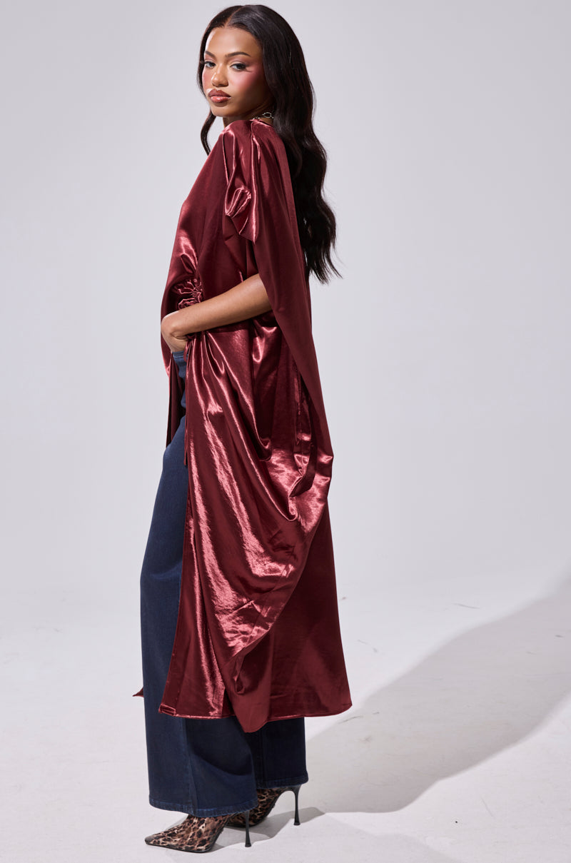 A woman, standing sideways against a plain light gray background, wears the shiny CLARA SIDE RUCHED BLOUSE in burgundy satin with an asymmetrical hemline, paired with dark blue wide-leg jeans and leopard print high-heeled boots. 3