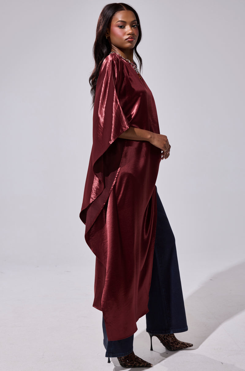 A woman in profile wears the CLARA SIDE RUCHED BLOUSE, a shiny maroon top with an asymmetrical hemline, paired with dark wide-leg pants and heeled leopard-print boots, against a plain light gray backdrop. 4