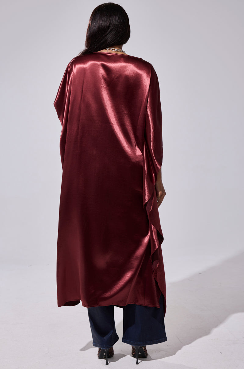 A person stands facing away, wearing blue pants, black shoes, and the oversized CLARA SIDE RUCHED BLOUSE under a long deep red satin cape against a plain light gray background. 5