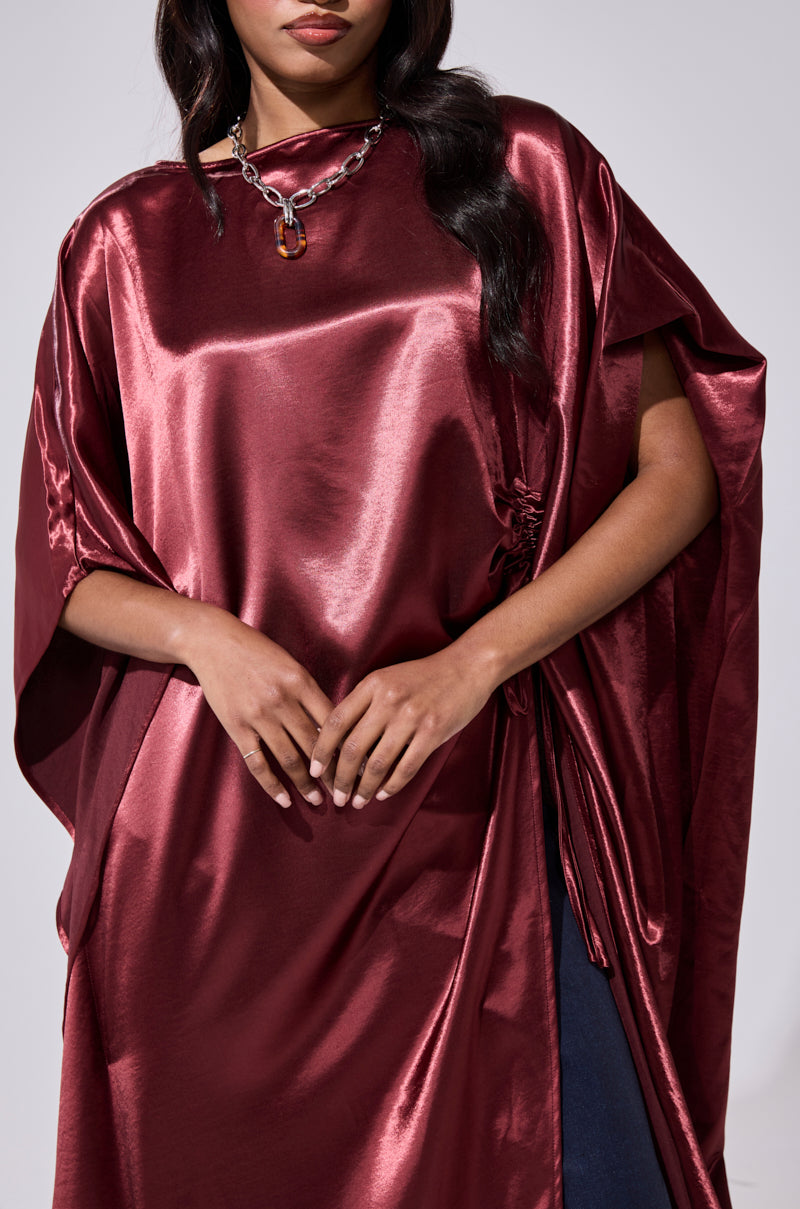 A woman wears the CLARA SIDE RUCHED BLOUSE, a shiny burgundy satin top with batwing sleeves and a silver chain necklace, standing with hands clasped at her waist. The photo is cropped at the shoulders and mid-thigh. 6