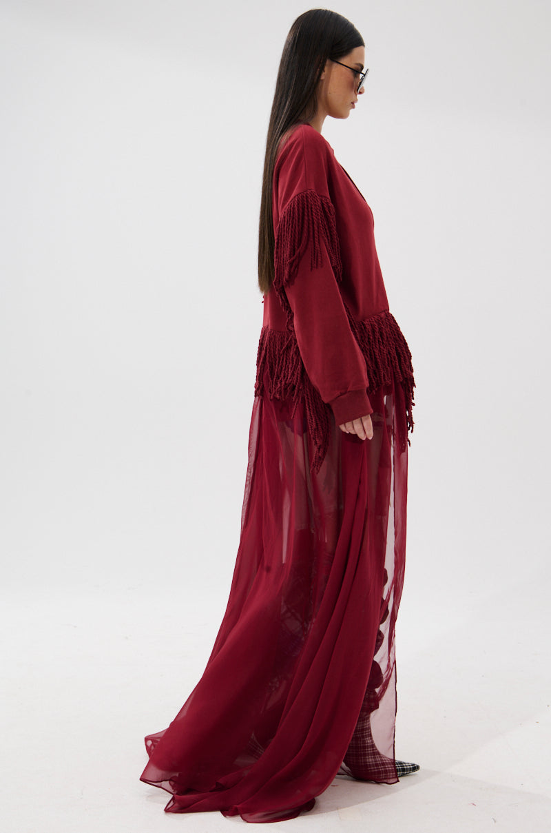 A woman with long dark hair and sunglasses stands in profile against a white background, wearing the GUESS WHO UNIQUE SWEATSHIRT—a deep red piece with elegant fringe trim and a sheer, flowing skirt.