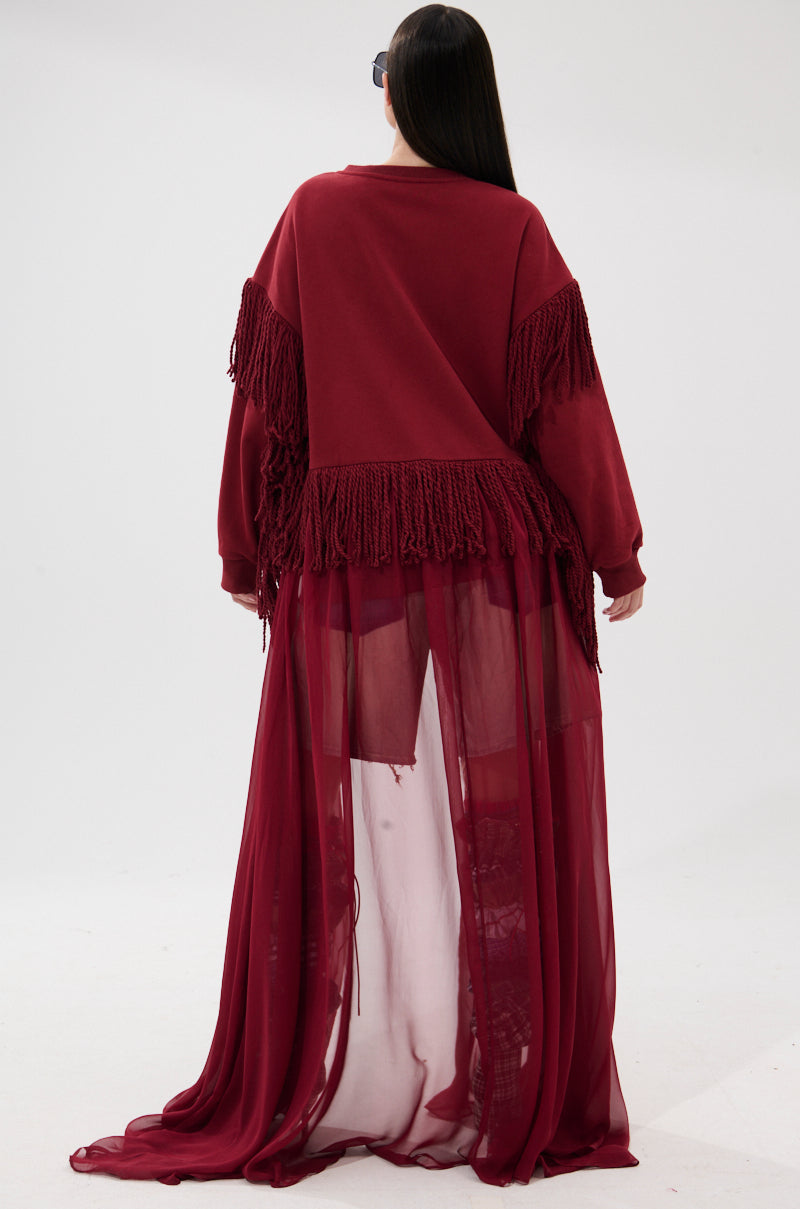 A person with long dark hair faces away, wearing the GUESS WHO UNIQUE SWEATSHIRT in burgundy with fringe trim and a sheer, floor-length skirt over shorts, set against a plain white background.
