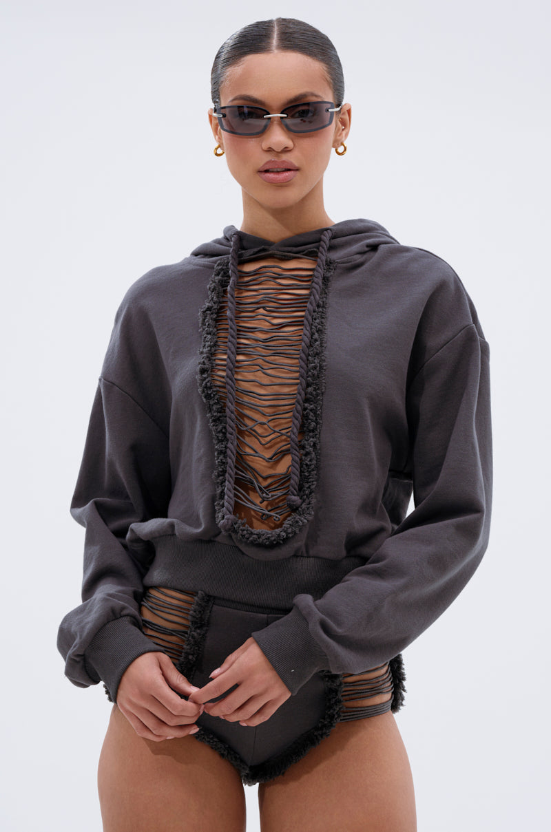 A woman in the STRESSED AND DISTRESSED SWEATSHIRT with fringe and lace-up front, paired with sunglasses, gold hoops, a tiger-striped top, and high-waisted bottoms stands against a plain light background. 0