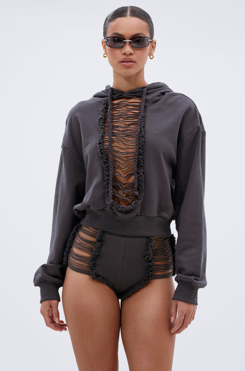 A woman in sunglasses and gold hoops models the STRESSED AND DISTRESSED SWEATHSIRT, a cropped hoodie with sheer ruched mesh panels and lace-up front, paired with matching high-waisted mesh-detailed shorts against a light backdrop. 1