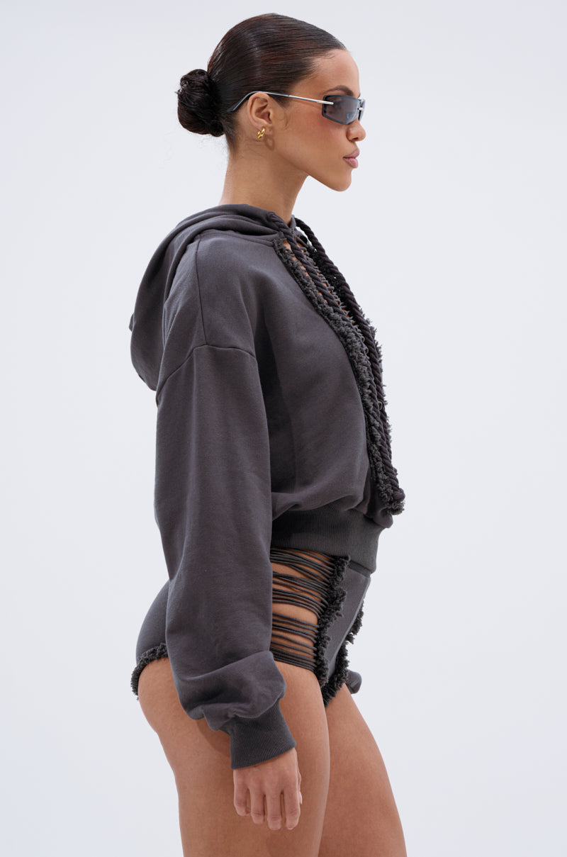 A woman in sunglasses and gold earrings models the STRESSED AND DISTRESSED SWEATHSIRT with black high-waisted shorts featuring side cutouts, standing in profile against a plain light background. 2