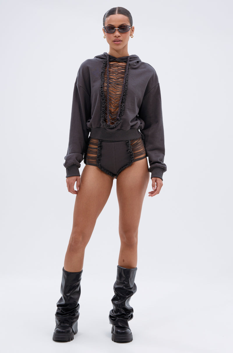 A woman wearing sunglasses, tall black boots, and the STRESSED AND DISTRESSED SWEATHSIRT—a dark cropped hoodie with frayed edges and lace-up front—plus matching high-waisted bottoms with sheer accents stands against a plain white background. 6