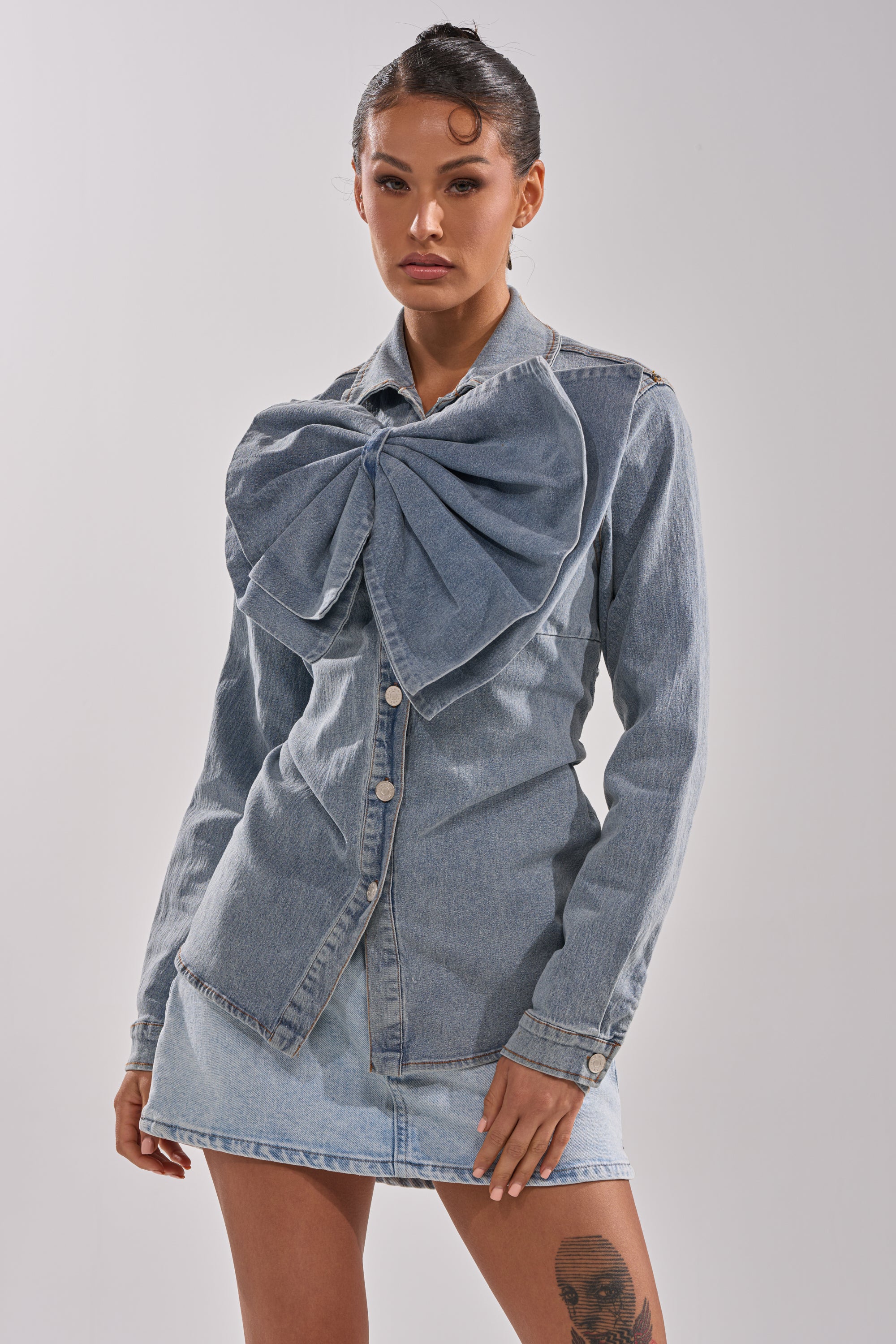 A woman wears the TWO-TONE BOW DENIM BUTTON DOWN SHIRT featuring an oversized denim bow and a matching mini skirt, standing against a plain background with her hair styled back and a thigh tattoo visible.