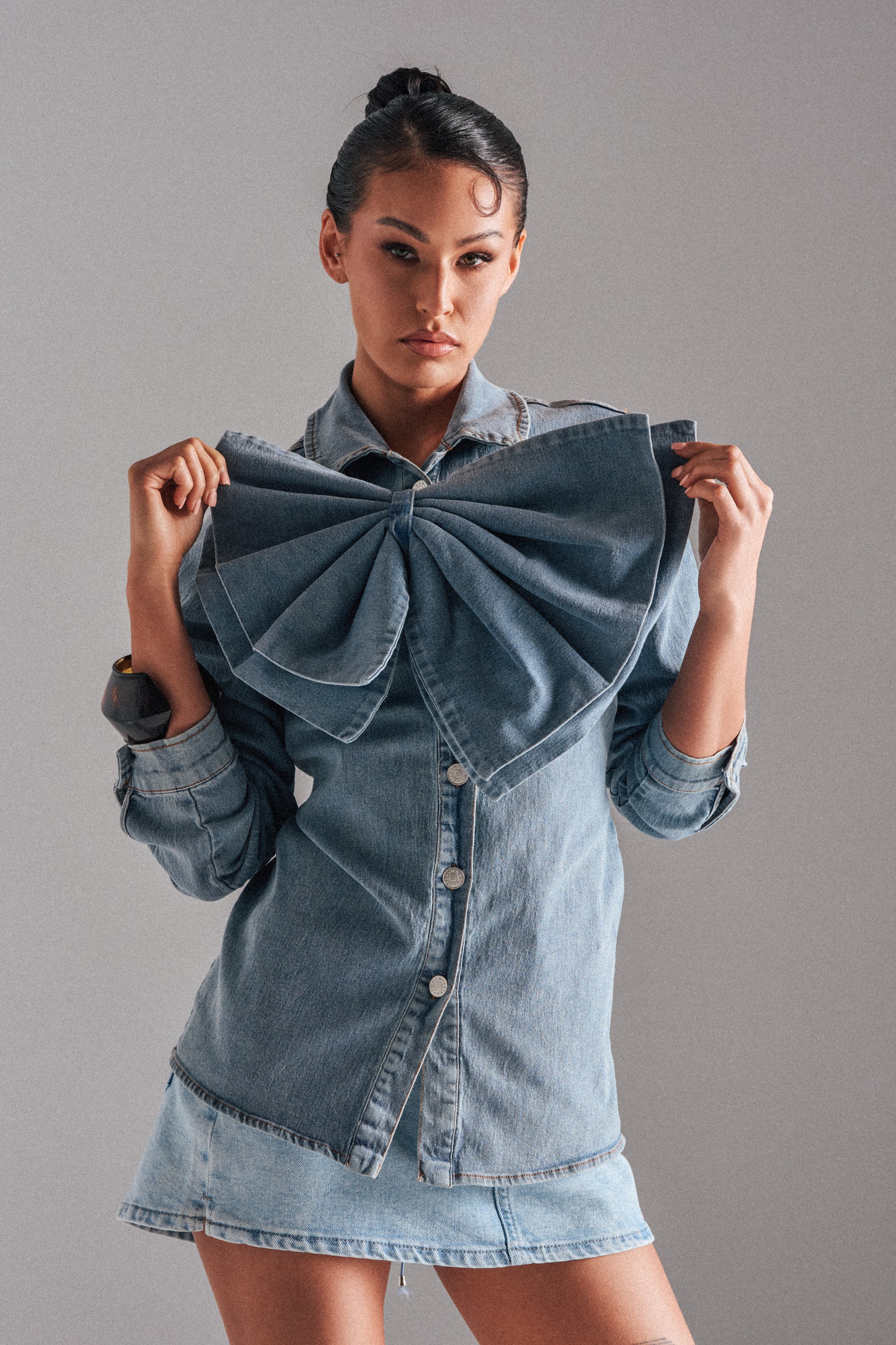 A woman wearing the TWO-TONE BOW DENIM BUTTON DOWN SHIRT with a large decorative bow on the chest and a light denim skirt poses against a gray background, holding the bow and looking confidently at the camera.