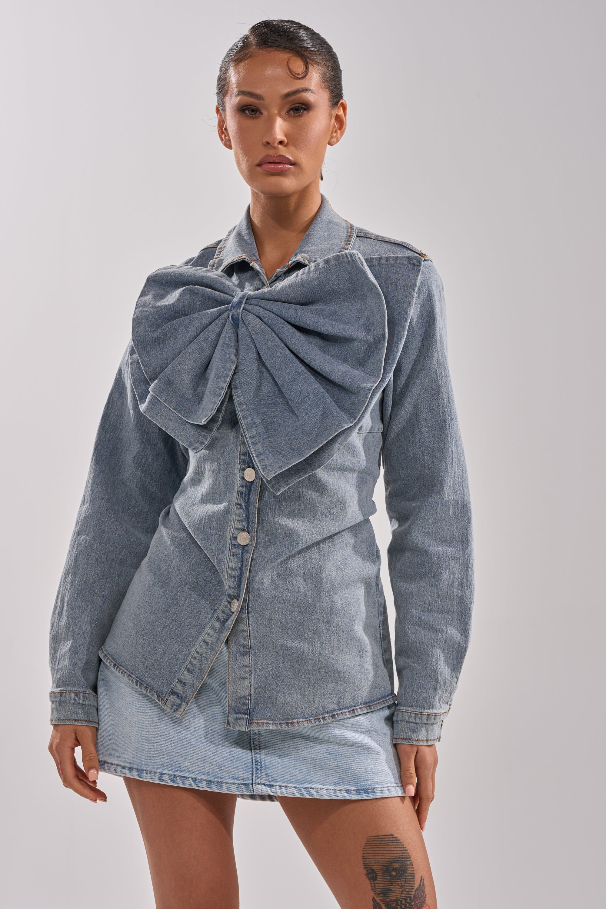 A woman styles her slicked-back hair and thigh tattoo with the TWO-TONE BOW DENIM BUTTON DOWN SHIRT, featuring a light blue oversized bow on the chest, paired with a matching mini skirt.
