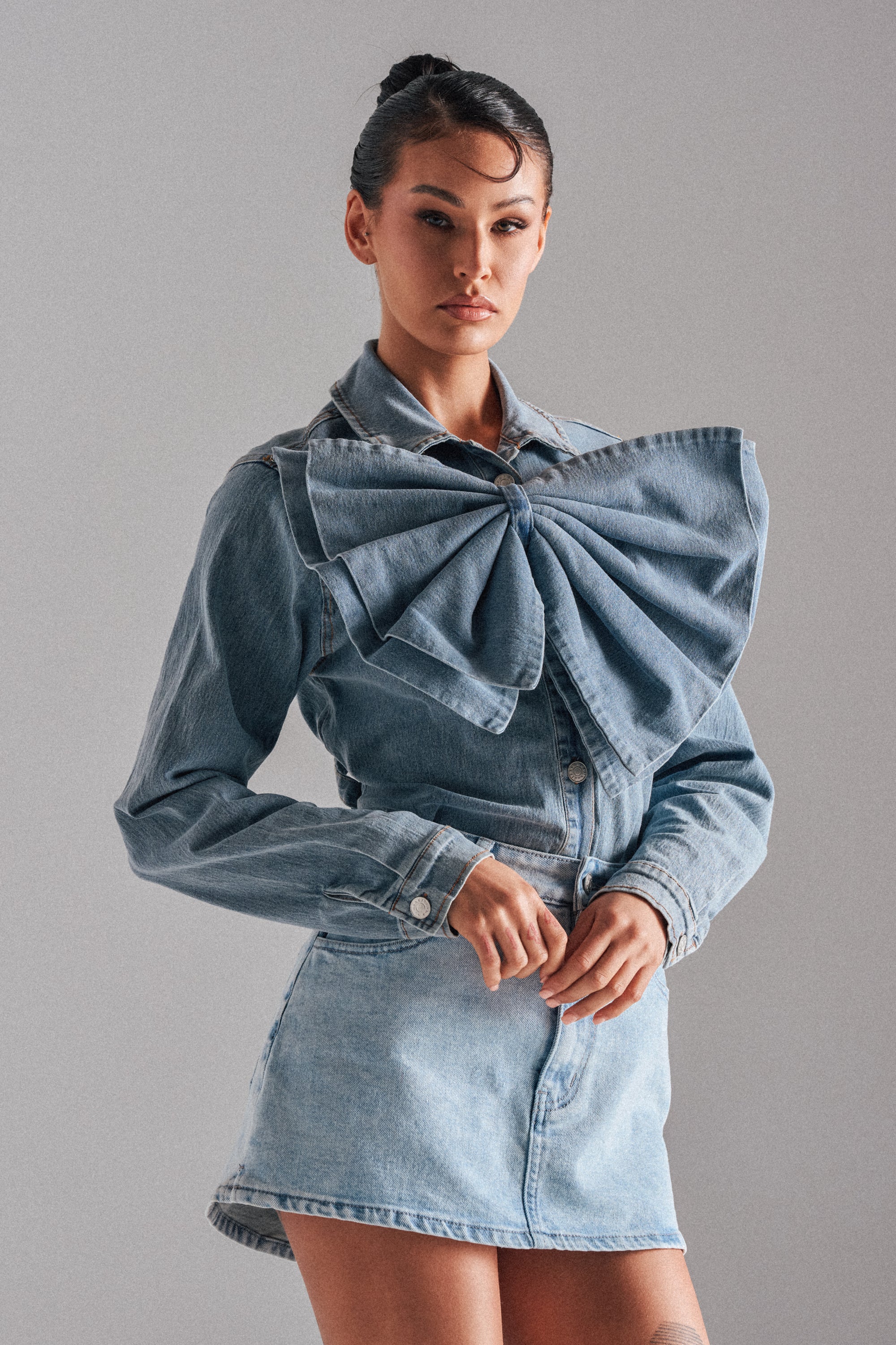 A woman wears the TWO-TONE BOW DENIM BUTTON DOWN SHIRT with an oversized bow and a matching mini skirt, standing against a plain gray background.