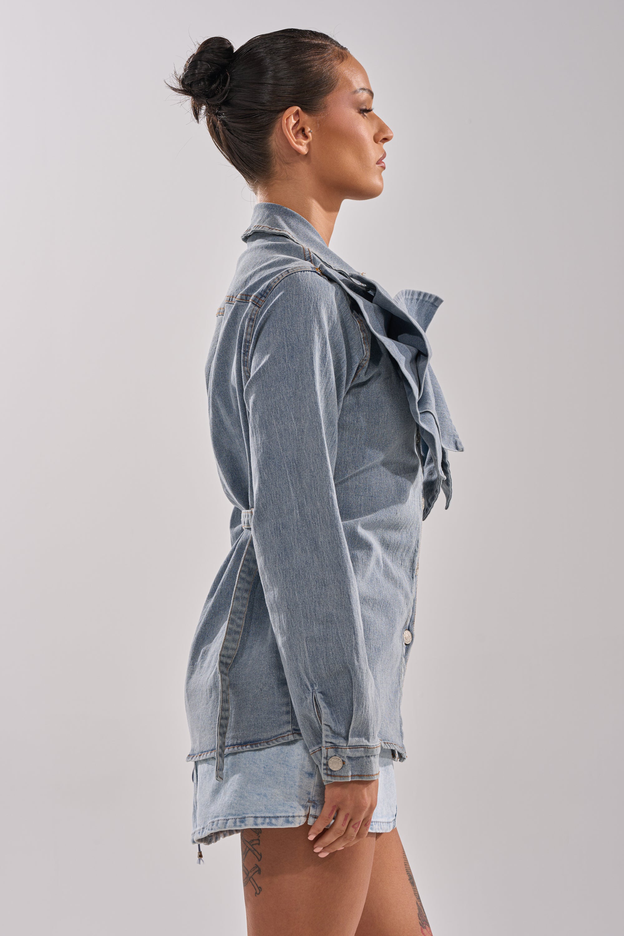 A woman with dark hair in a bun stands in profile wearing the TWO-TONE BOW DENIM BUTTON DOWN SHIRT and matching denim shorts against a plain grey background.