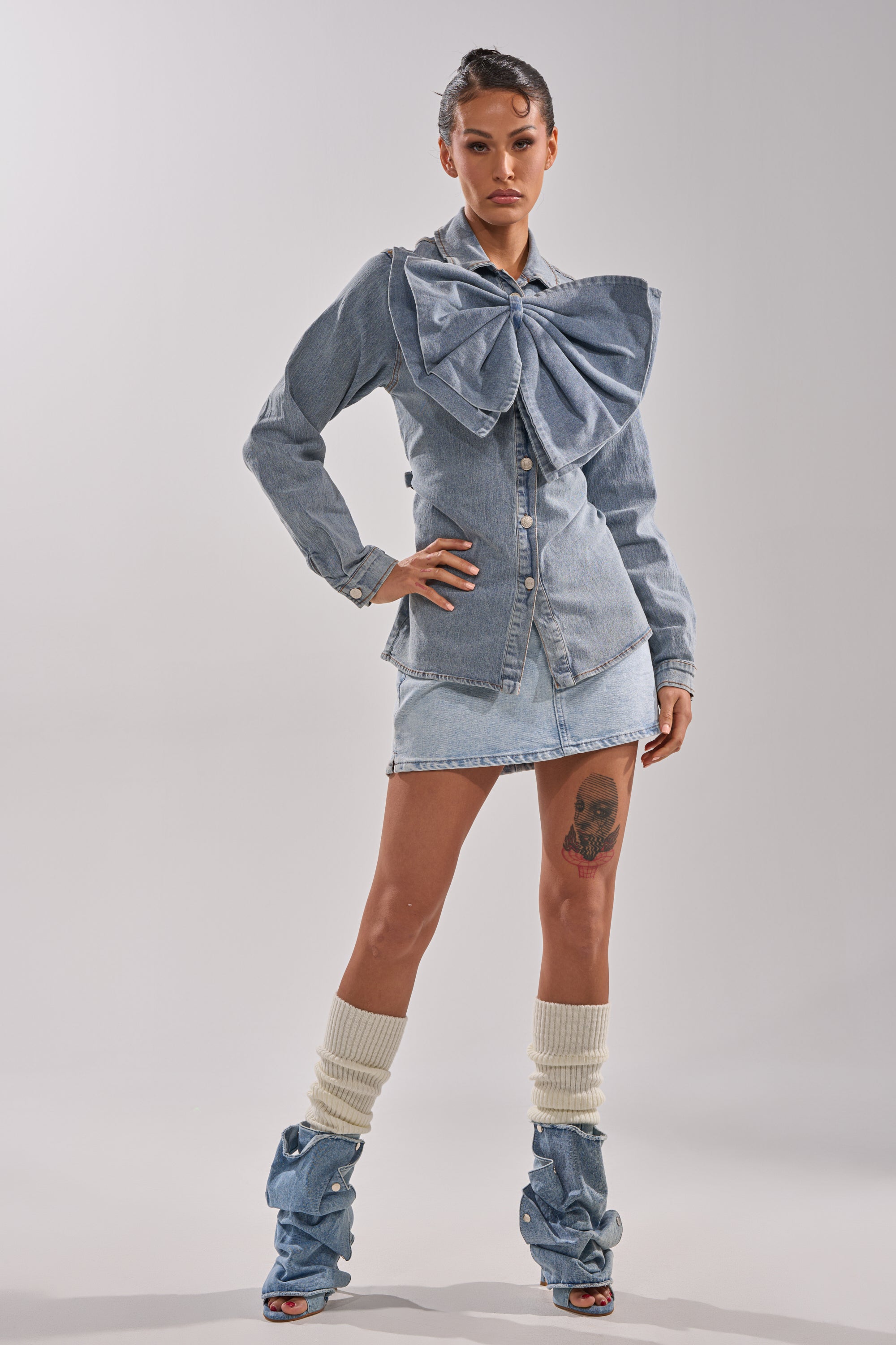 A woman models the TWO-TONE BOW DENIM BUTTON DOWN SHIRT with a matching denim mini skirt and slouchy boots, highlighting her thigh tattoo against a plain background.