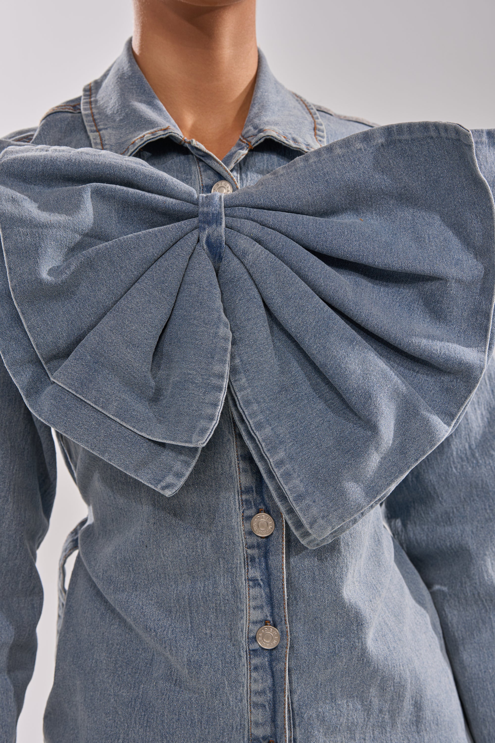 Someone wears the TWO-TONE BOW DENIM BUTTON DOWN SHIRT, a light-wash cotton denim shirt featuring large buttons and an oversized denim bow on the chest, set against a plain, light background.