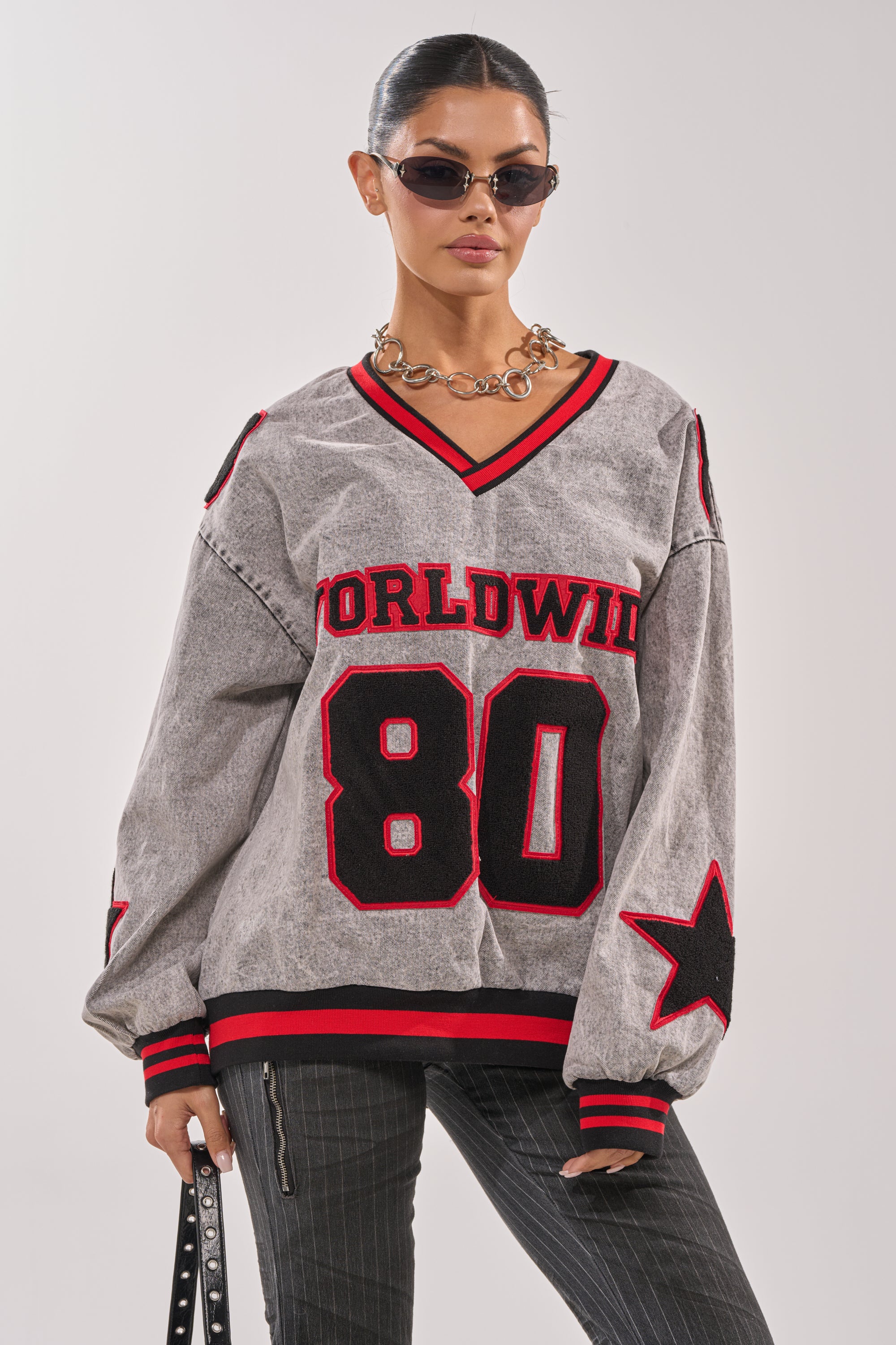 A woman wears sunglasses, a chunky chain necklace, a small black bag, and the IN MY OWN WORLD DENIM SWEATSHIRT—a gray collegiate style with "WORLDWIDE" and "80" in red/black letters and star embroidered patches on the sleeves. 0