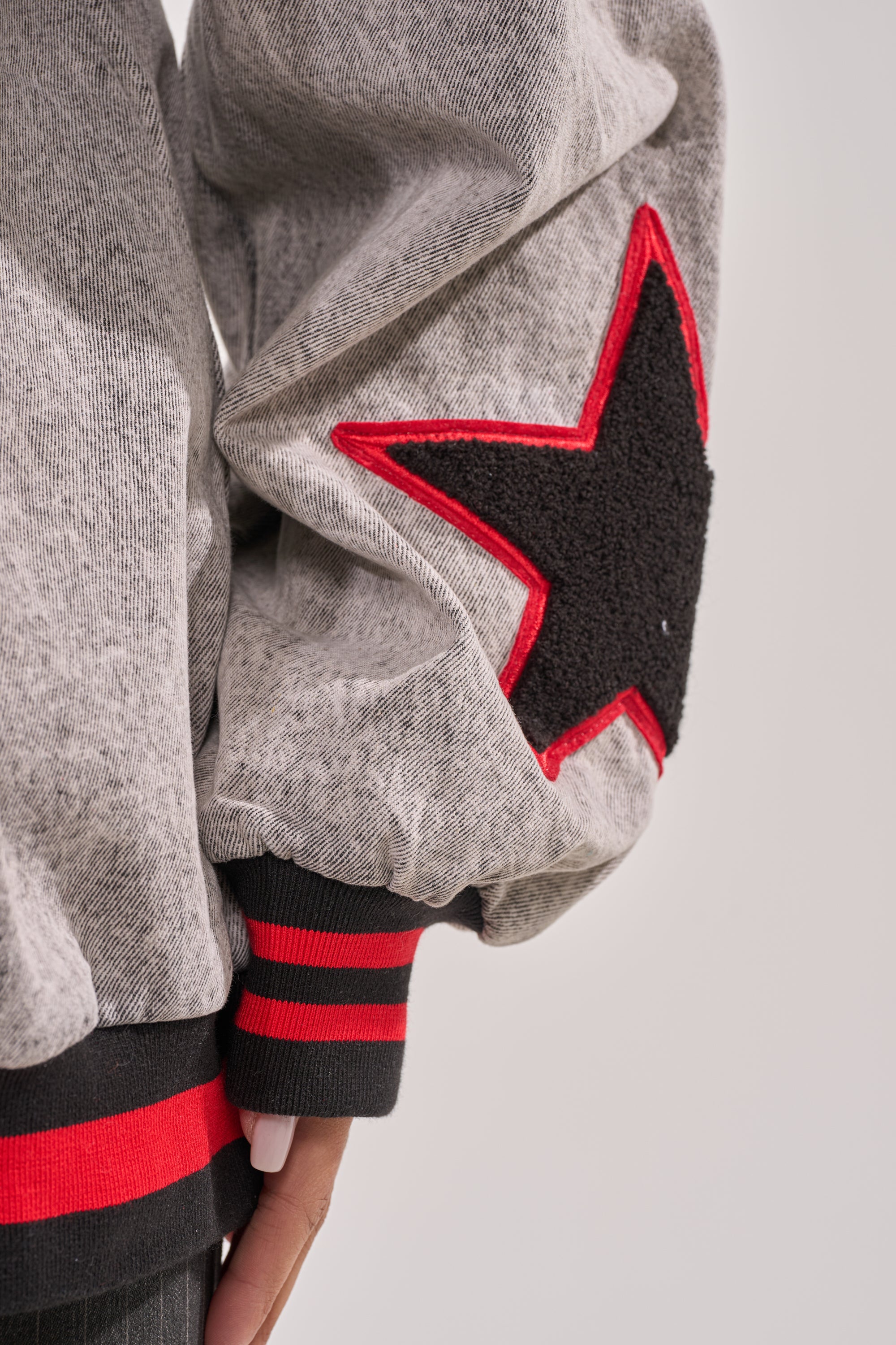 A close-up of the IN MY OWN WORLD DENIM SWEATSHIRT’s gray sleeve shows a large black star patch with red outline. The ribbed cuff features black and red stripes, enhancing its collegiate vibe. A hand is partially visible at the bottom. 9