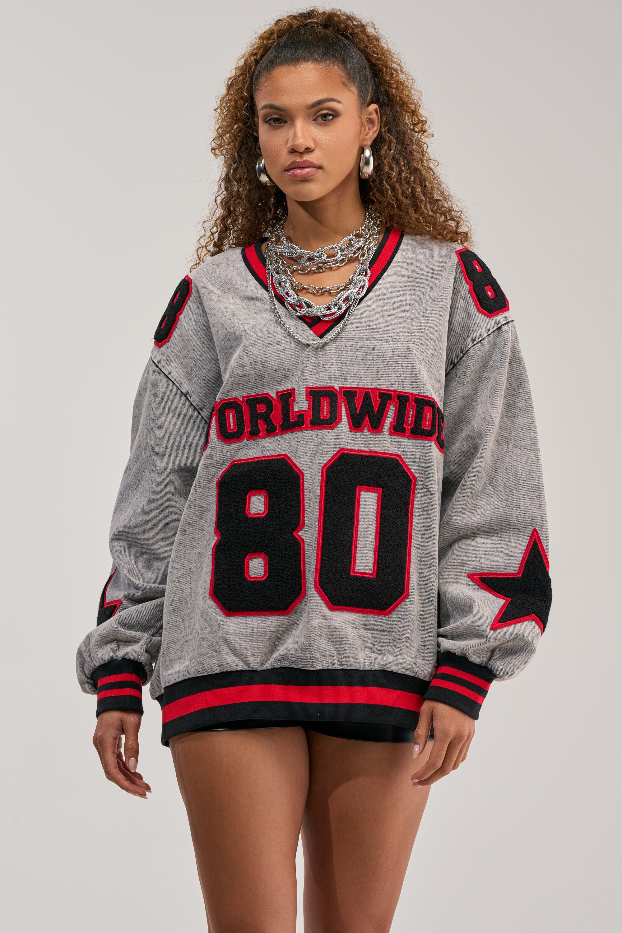 A woman models the IN MY OWN WORLD DENIM SWEATSHIRT—an oversized grey streetwear piece with "WORLDWIDE 80" in red and black, striped sleeves, a sporty V-neckline, and a black star detail—against a plain background.