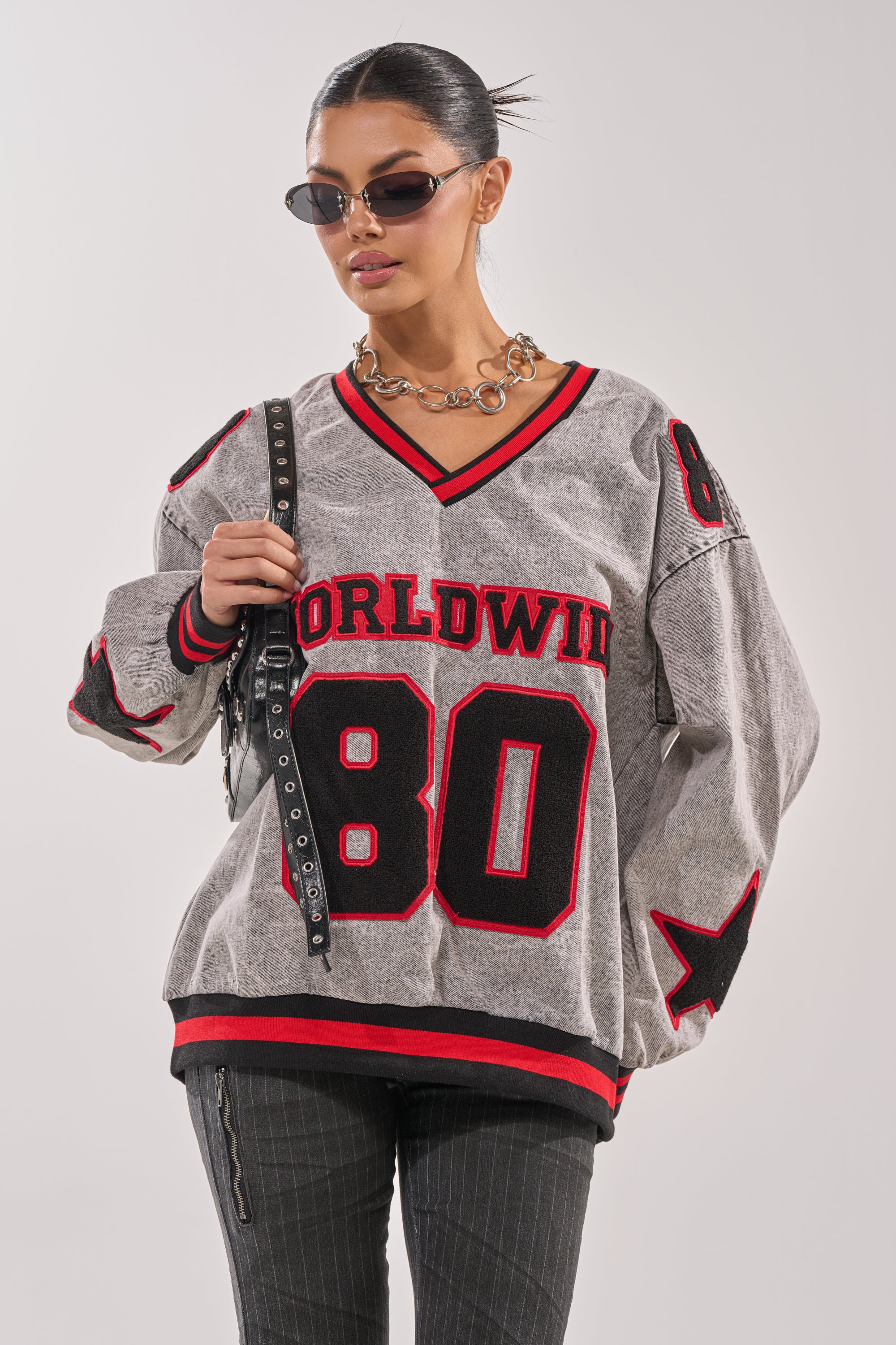 A woman channels collegiate style wearing sunglasses, a chunky chain necklace, dark pinstriped pants, and the IN MY OWN WORLD DENIM SWEATSHIRT with "WORLDWIDE 80" and red-black accents, standing against a plain light background. 1