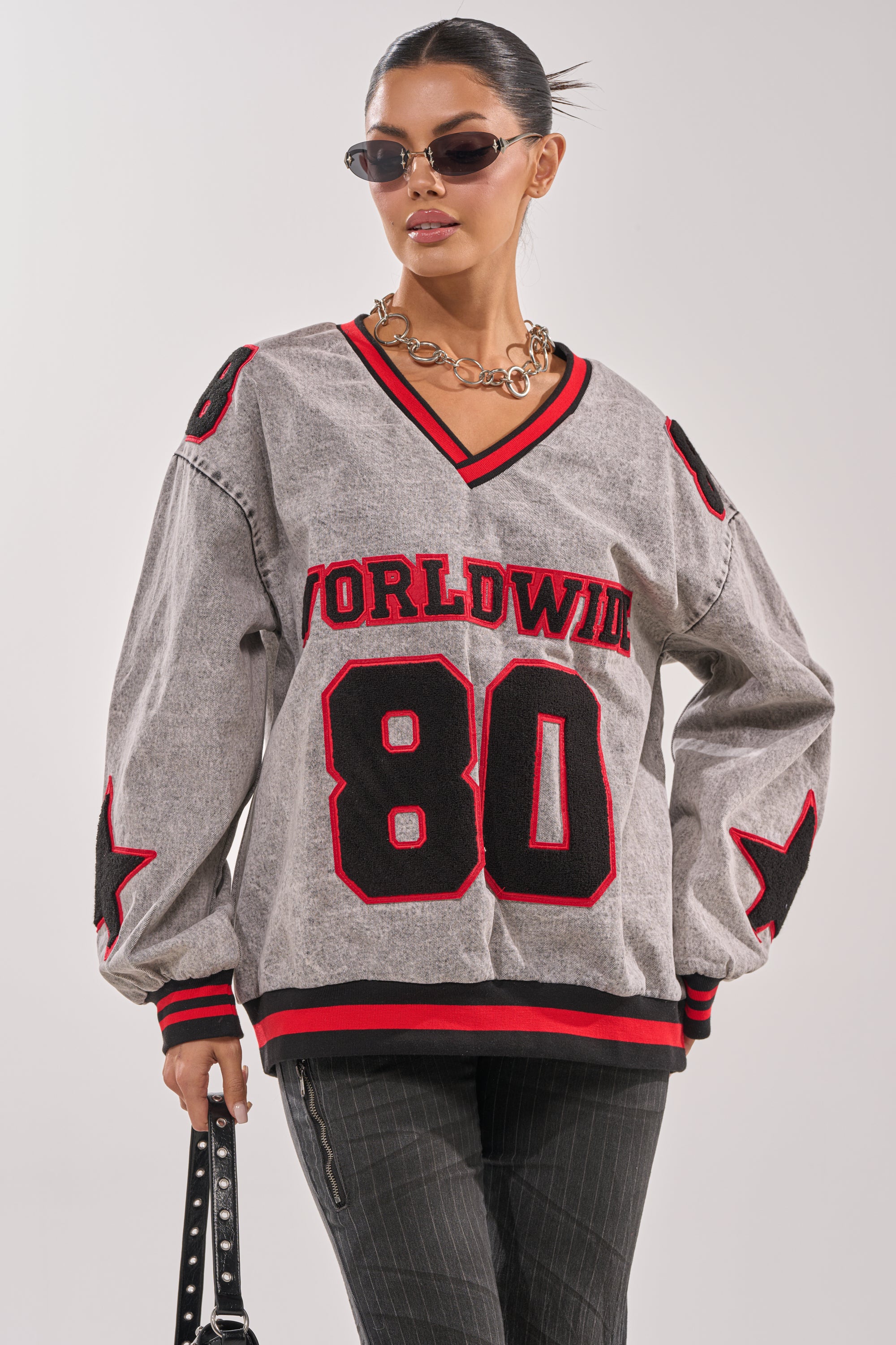 A woman in sunglasses and a chunky chain necklace poses in the loose gray IN MY OWN WORLD DENIM SWEATSHIRT with "WORLDWIDE 80" and patch details, paired with pinstriped pants and a black bag. 2