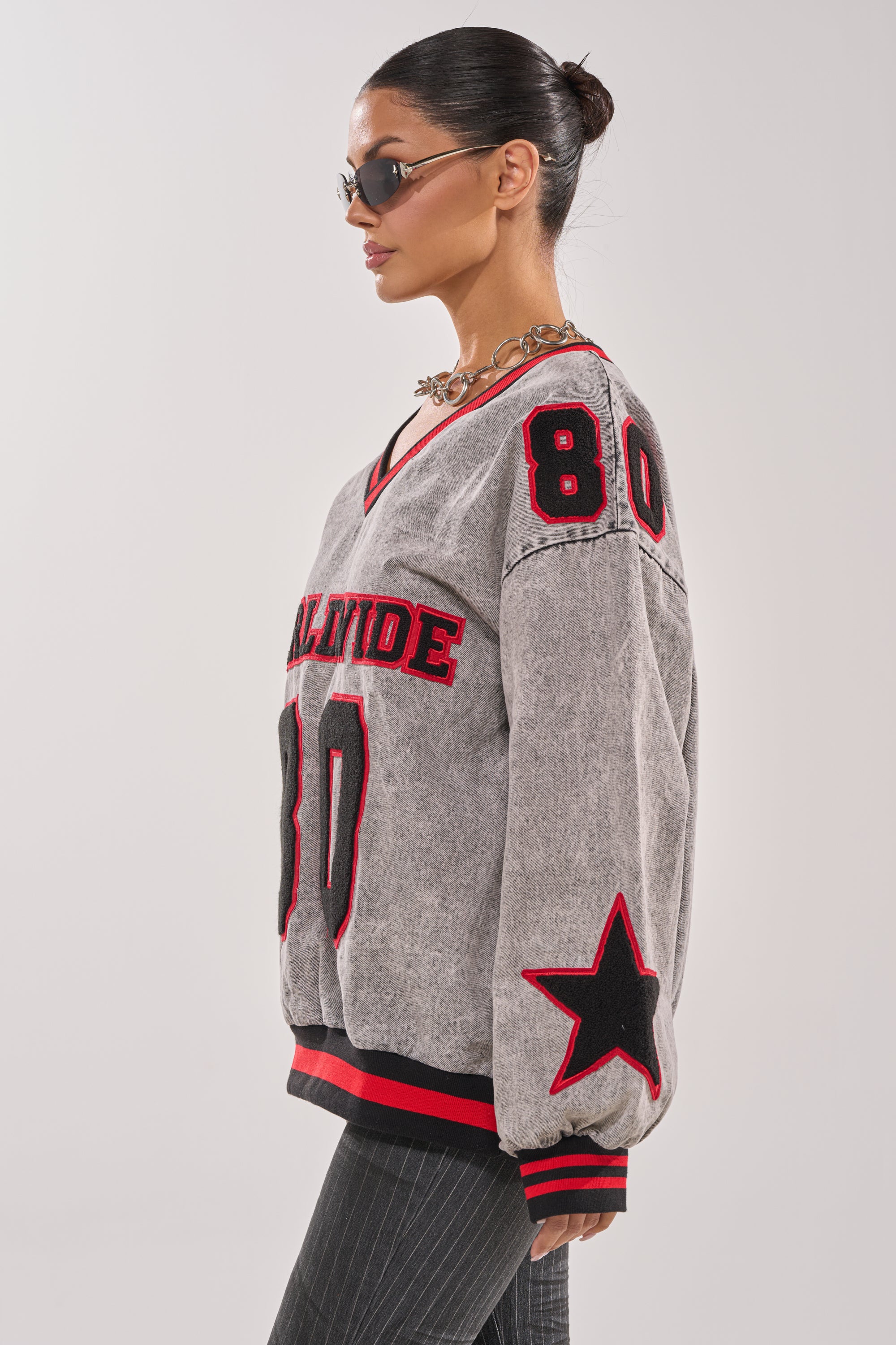 A woman in sunglasses and a chunky chain necklace models the IN MY OWN WORLD DENIM SWEATSHIRT, featuring red and black accents, "ALTITUDE" text, the number 80, and an embroidered sleeve patch, posing in profile against a plain background. 3