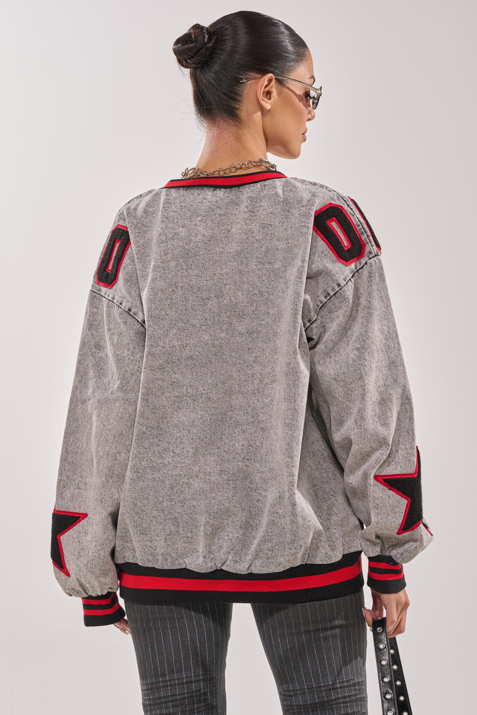 A woman with dark hair in a bun, facing away from the camera, wears the IN MY OWN WORLD DENIM SWEATSHIRT—an oversized varsity-style jacket with embroidered patches and stars—while holding a studded bag. 4