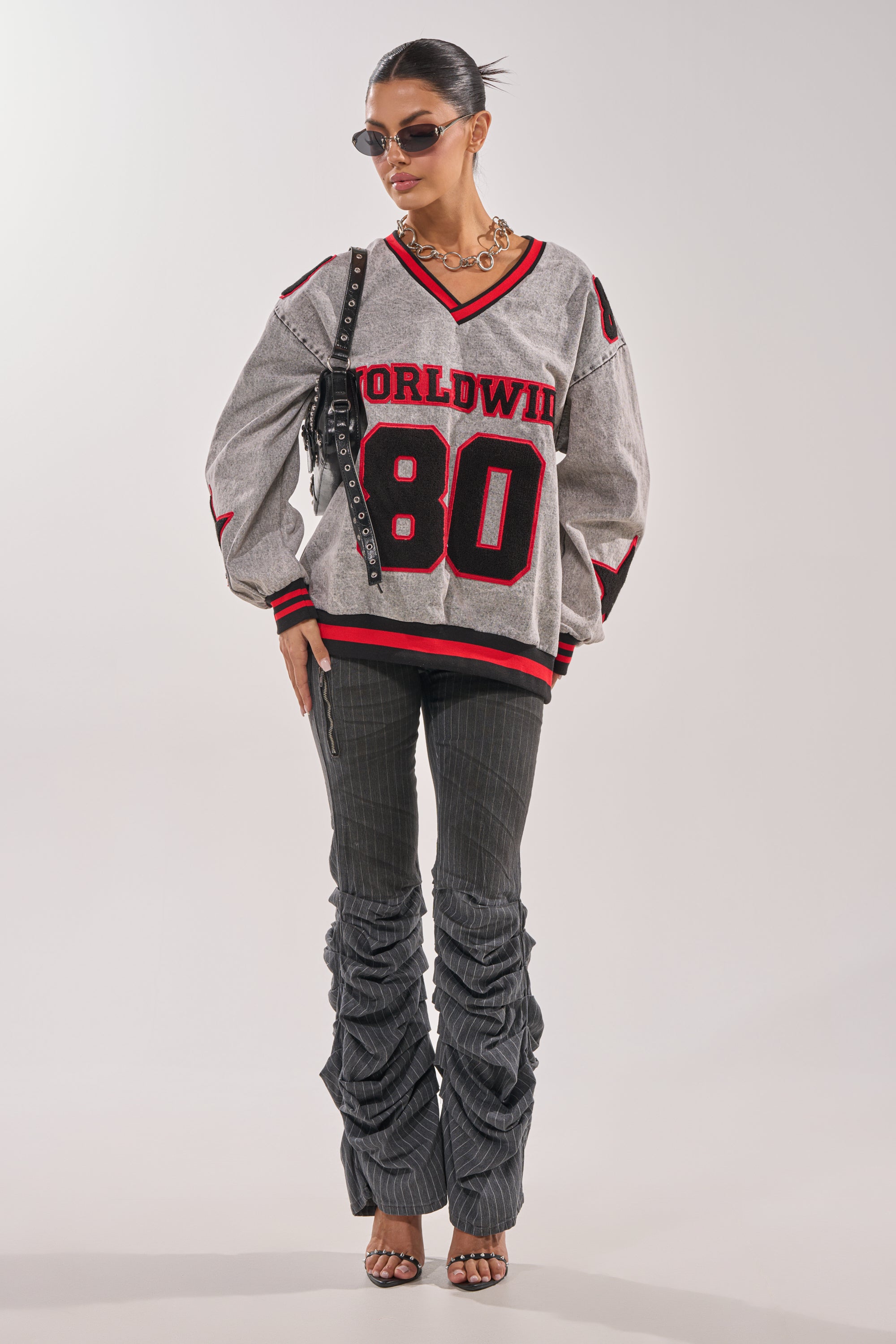 A woman in sunglasses, layered necklaces, dark pinstripe ruched pants, strappy heels, a black shoulder bag, and the gray IN MY OWN WORLD DENIM SWEATSHIRT with collegiate details stands confidently against a plain background. 5