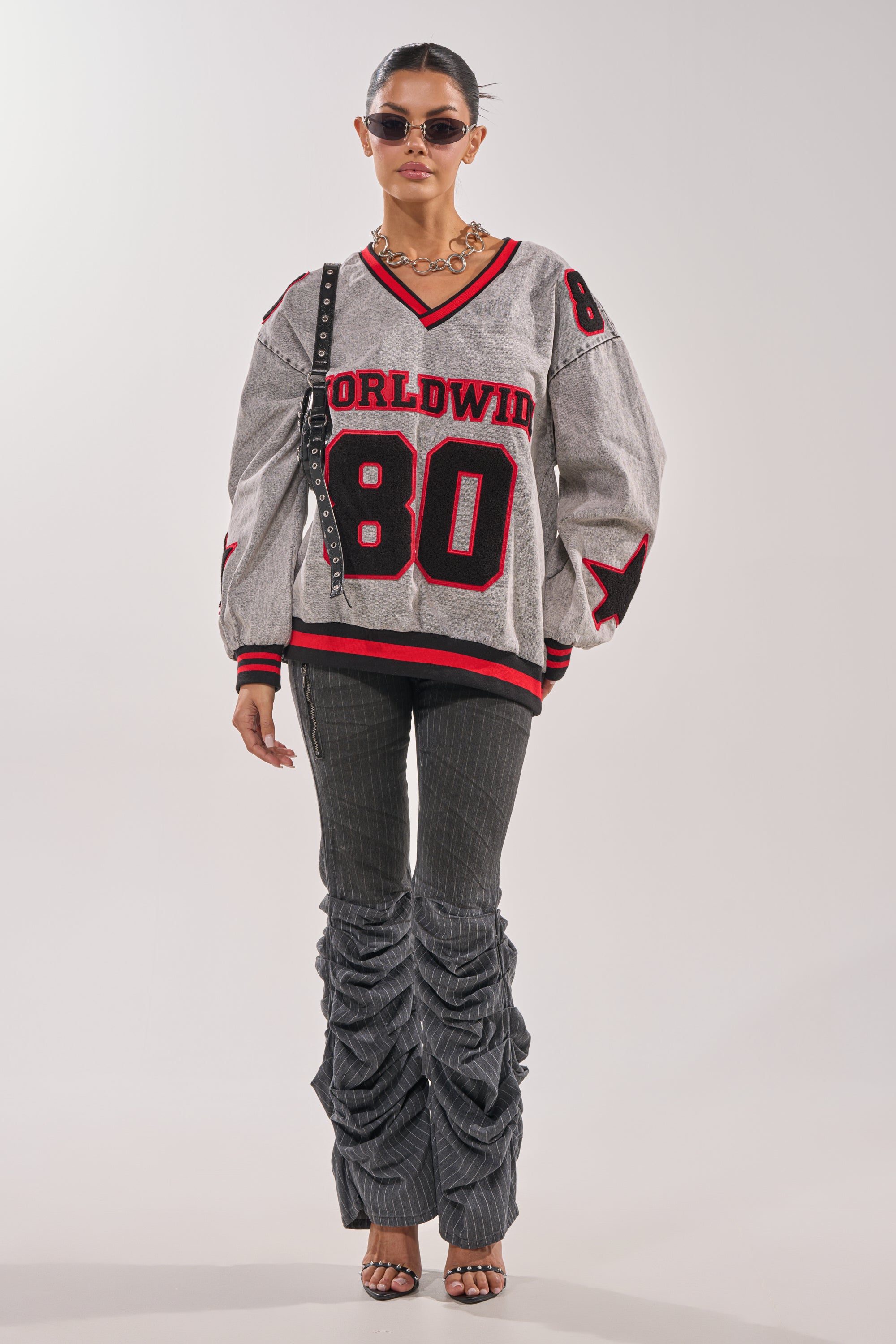 A woman wearing sunglasses, a chunky necklace, the IN MY OWN WORLD DENIM SWEATSHIRT with “Worldwide 80” and patch details, striped ruched pants, heels, and a studded crossbody bag stands against a plain background. 6