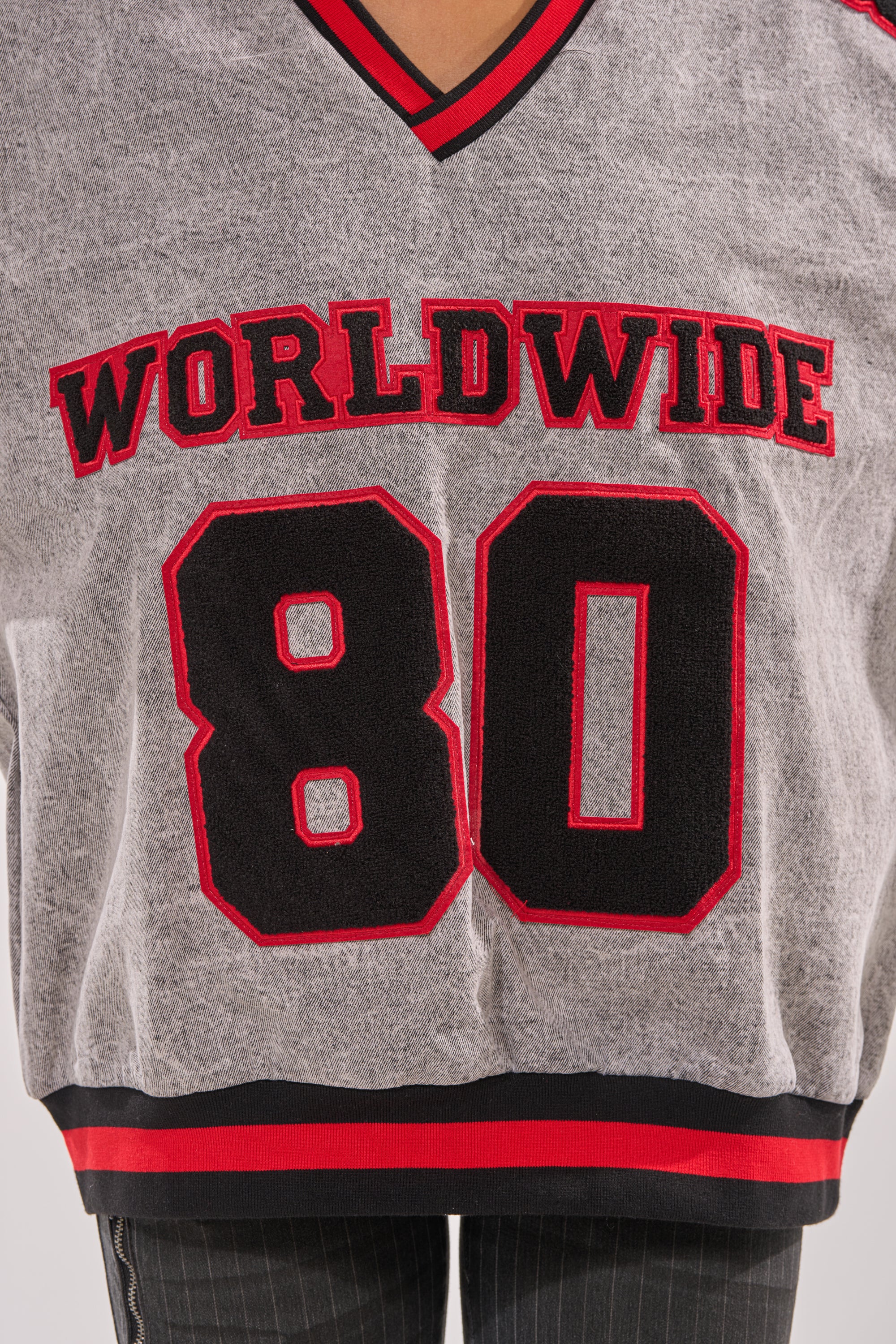 A close-up of the IN MY OWN WORLD DENIM SWEATSHIRT, a gray collegiate style with red and black trim, "WORLDWIDE," the number "80," and bold embroidered patches on the front. 8