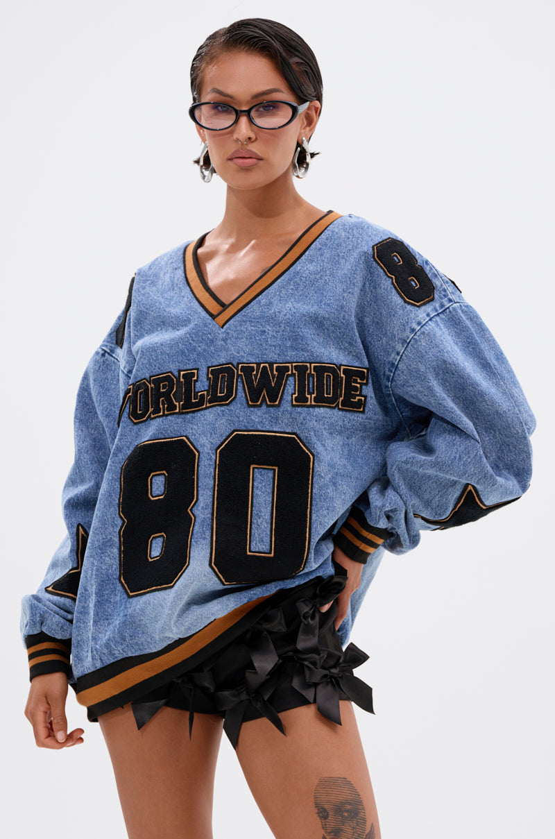 A person in cat-eye glasses, short hair, and black ruffled shorts confidently poses against a plain background wearing the ALL AROUND THE WORLD DENIM SWEATSHIRT with "WORLDWIDE 80" on front, revealing a thigh tattoo.
