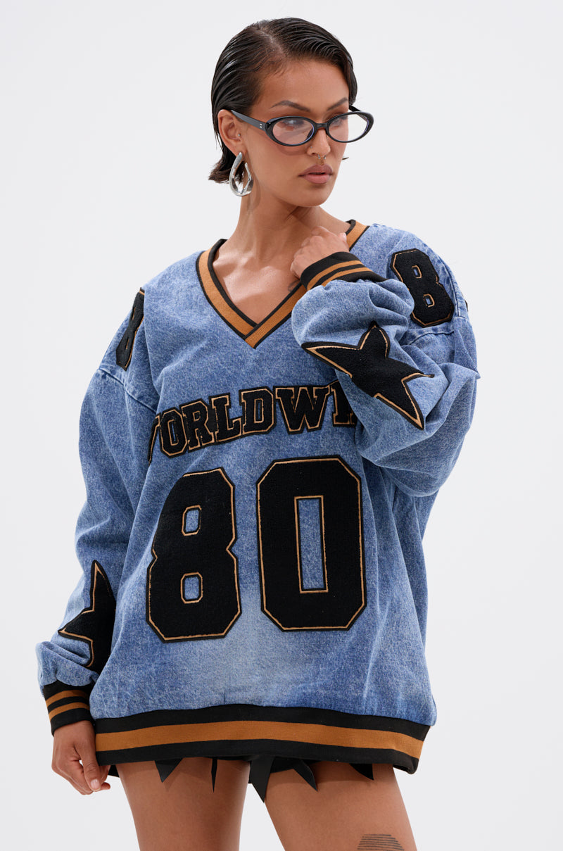 Someone wearing glasses and the ALL AROUND THE WORLD DENIM SWEATSHIRT, featuring "WORLDWIDE" and the number 80 in black letters with black and gold star patches, stands against a plain light background.