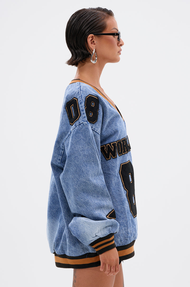 A person with short dark hair, sunglasses, and hoop earrings wears the ALL AROUND THE WORLD DENIM SWEATSHIRT—an oversized blue varsity-style top with black and gold patches featuring "8" and "WORLD.